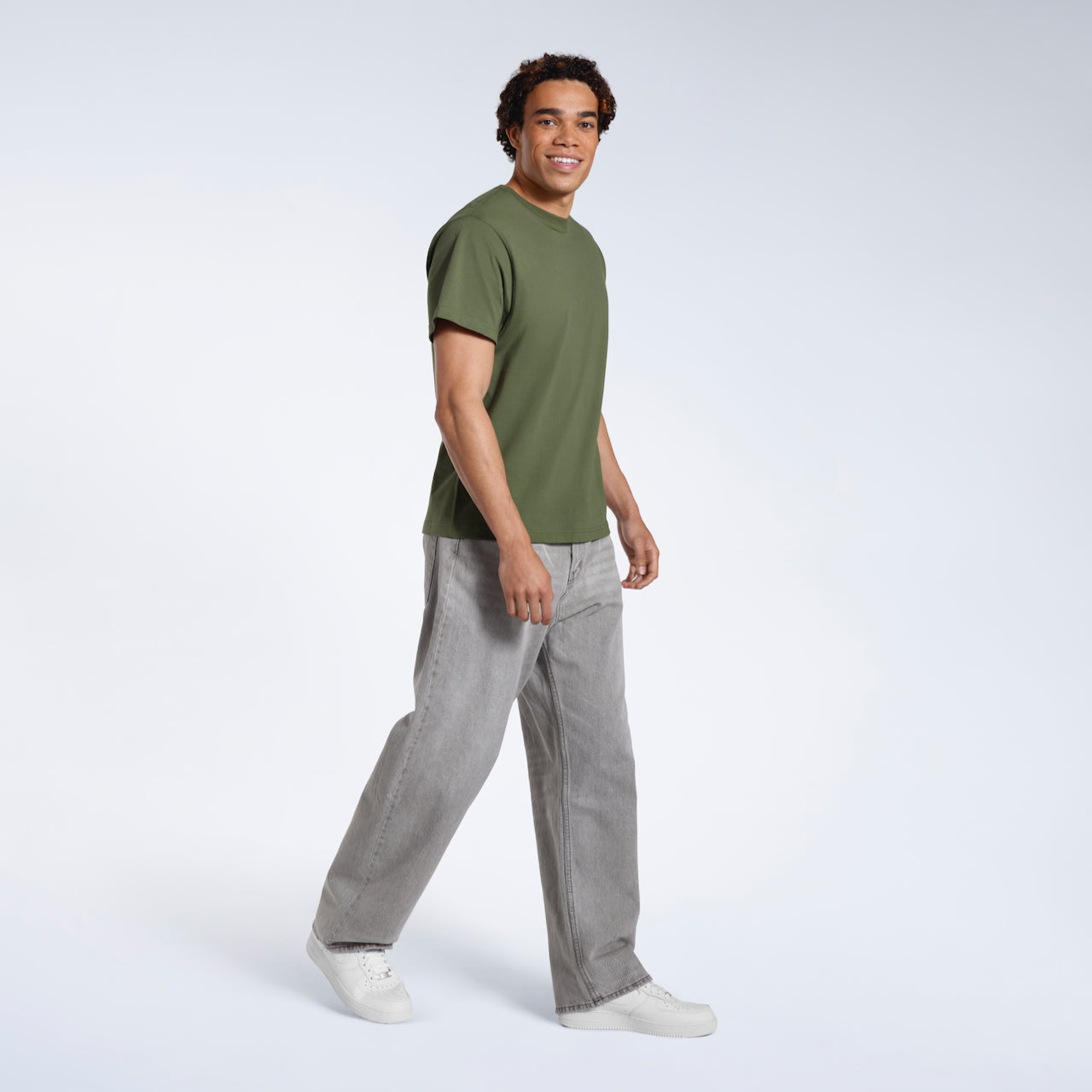 A full body photo of a man looking into the camera smiling, modelling a plain khaki green short sleeve organic cotton t shirt.