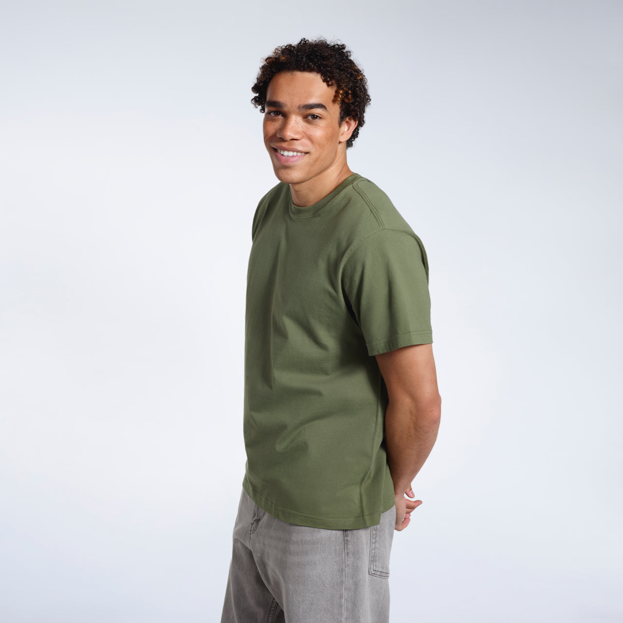 A man smiling into the camera with his hands behind his back, modelling a plain khaki green short sleeve organic cotton t shirt.