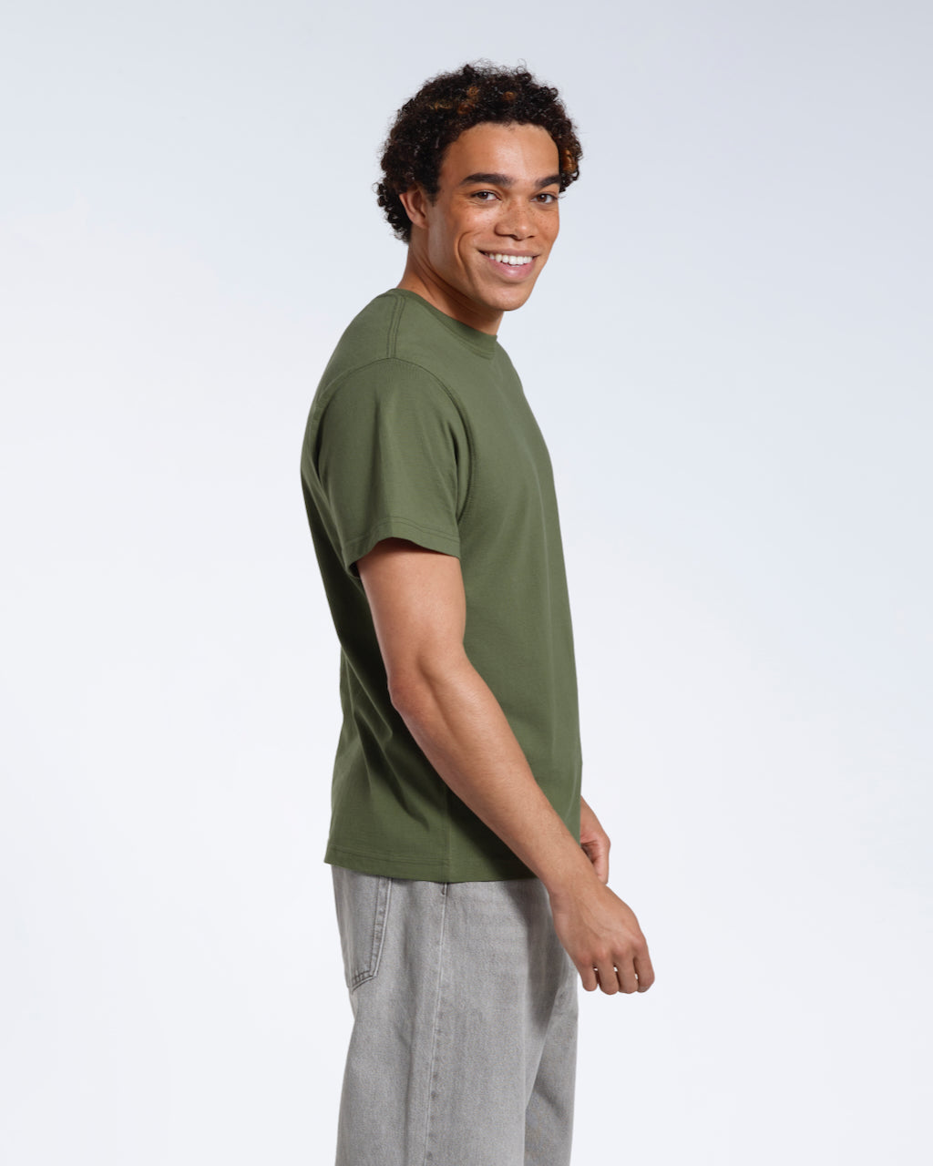 A man facing side on with his head turned towards the camera smiling, modelling a plain khaki green short sleeve organic cotton t shirt.