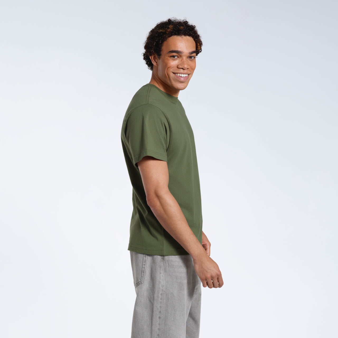 A man facing side on with his head turned towards the camera smiling, modelling a plain khaki green short sleeve organic cotton t shirt.