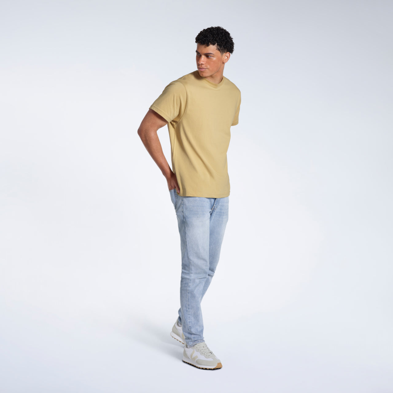 A full body photo of a man looking to the side with his hands in his back pockets, modelling a plain light khaki short sleeve organic cotton t shirt.