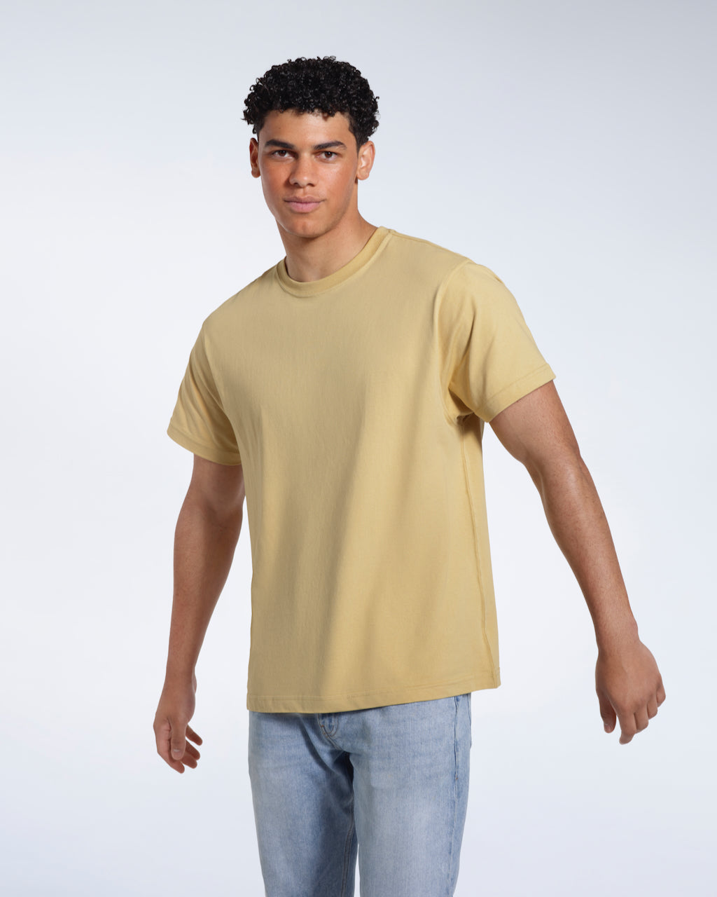 A man looking into the camera with his arms slightly raised, modelling a plain light khaki short sleeve organic cotton t shirt.