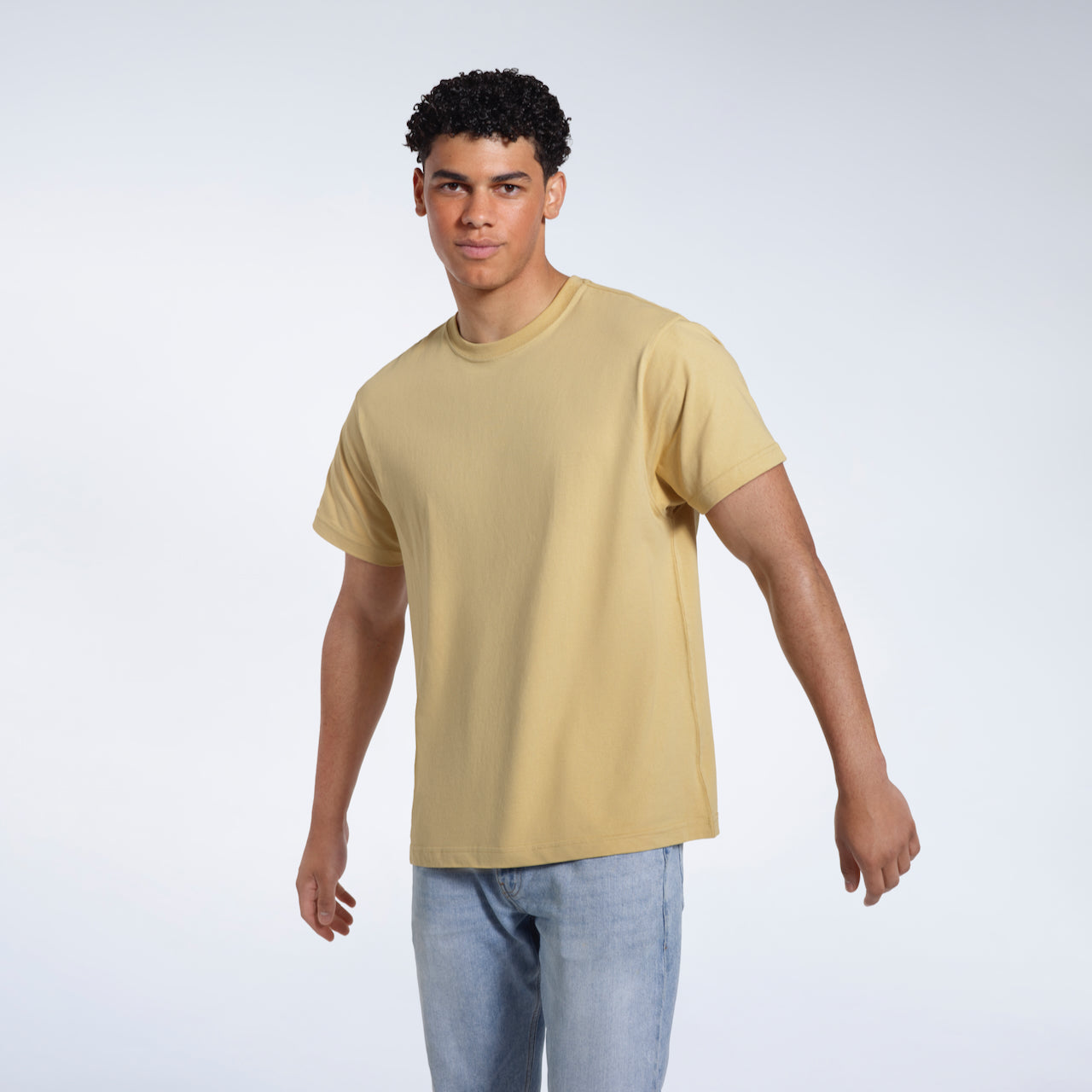 A man looking into the camera with his arms slightly raised, modelling a plain light khaki short sleeve organic cotton t shirt.