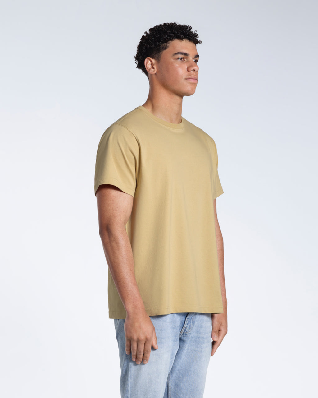 A man at an angle with his arms by his sides, modelling a plain light khaki short sleeve organic cotton t shirt.