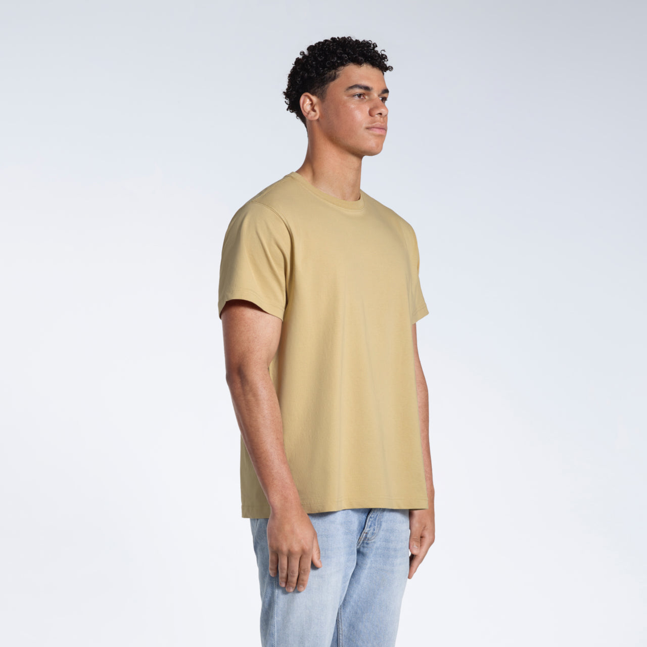 A man at an angle with his arms by his sides, modelling a plain light khaki short sleeve organic cotton t shirt.