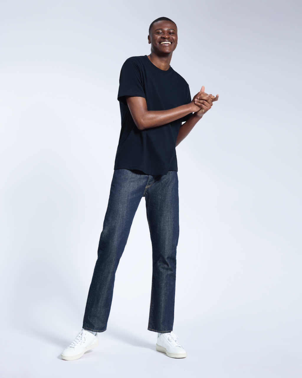A full body photo of a man smiling with his hands together, modelling a plain navy short sleeve organic cotton t shirt.
