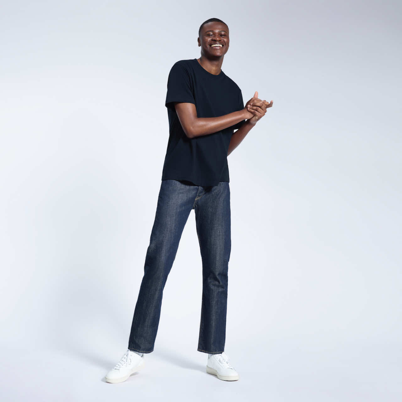 A full body photo of a man smiling with his hands together, modelling a plain navy short sleeve organic cotton t shirt.
