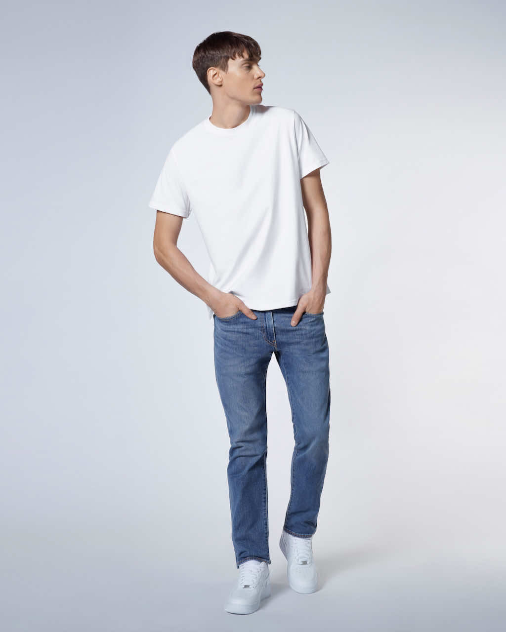 A full body photo of a man looking to the side with his hands in his jeans pockets, modelling a plain white short sleeve organic cotton t shirt.