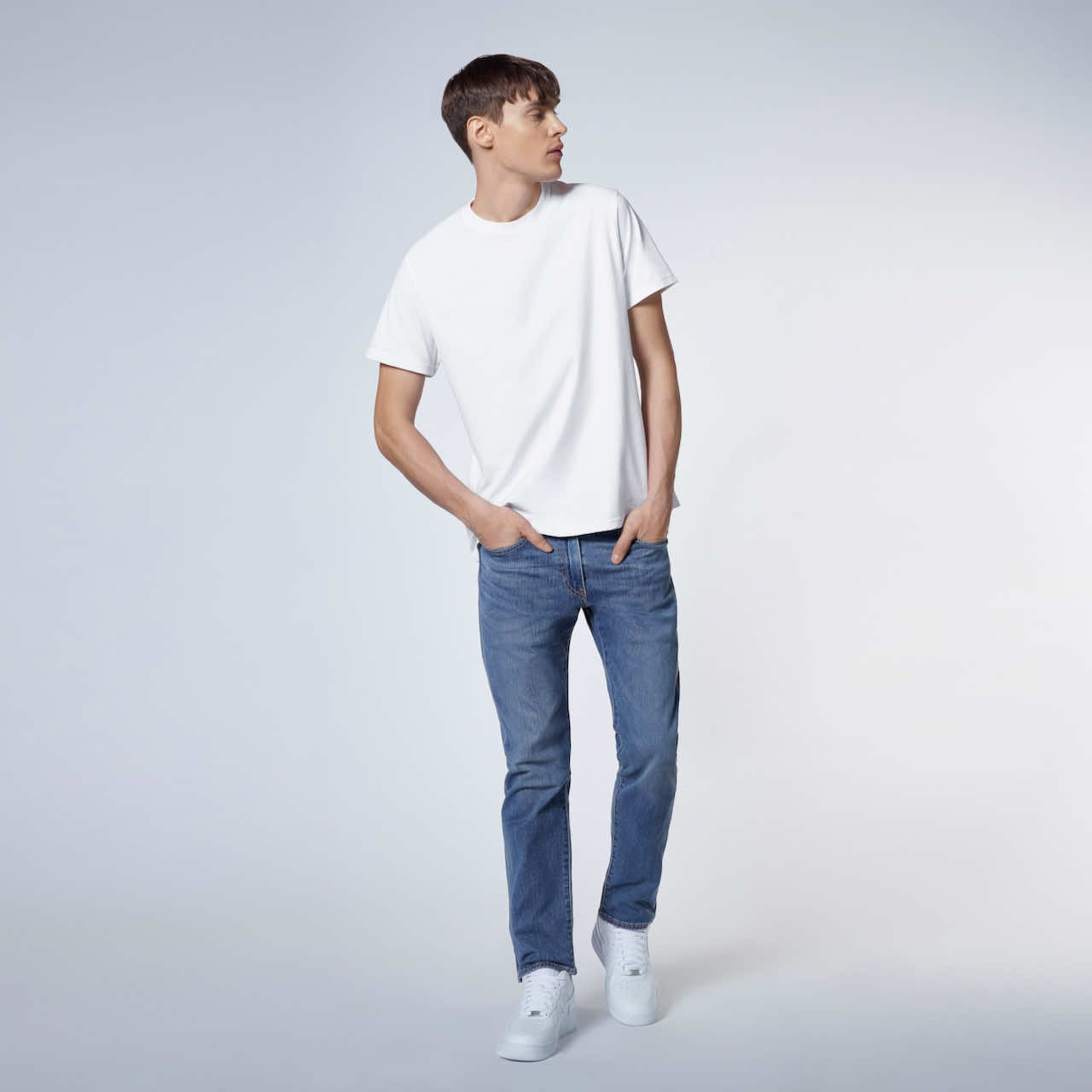 A full body photo of a man looking to the side with his hands in his jeans pockets, modelling a plain white short sleeve organic cotton t shirt.