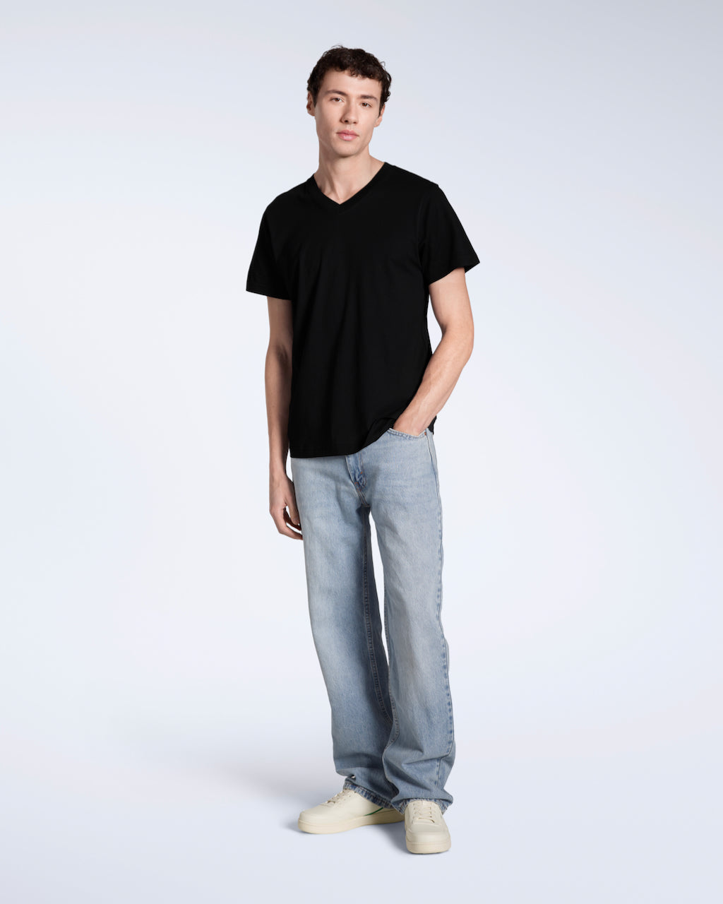 A full body photo of a man smiling looking away from the camera, modelling a plain black, organic cotton v neck t shirt with a pair of blue jeans.