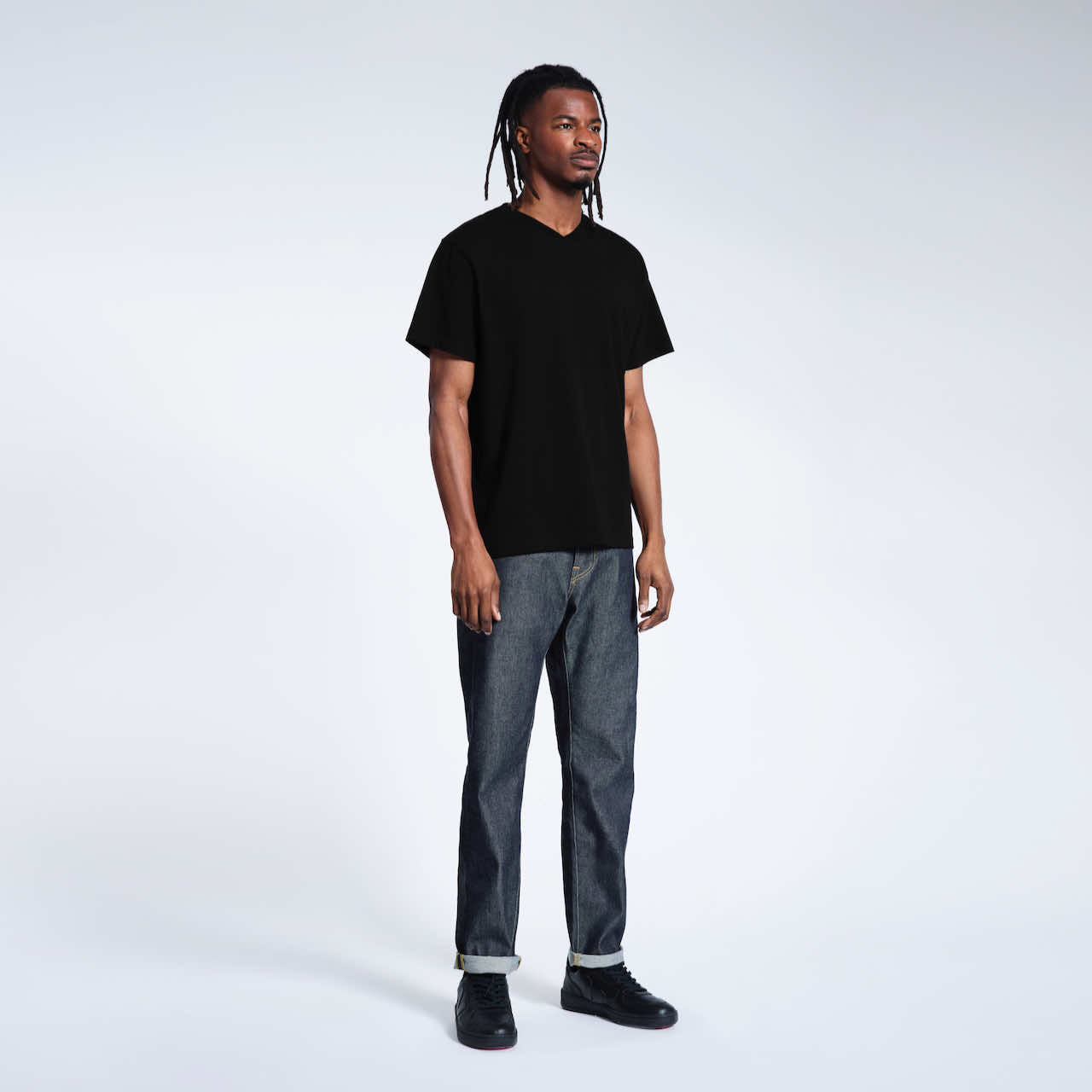 A full body photo of a man looking away from the camera, modelling a plain black organic cotton v neck t shirt.