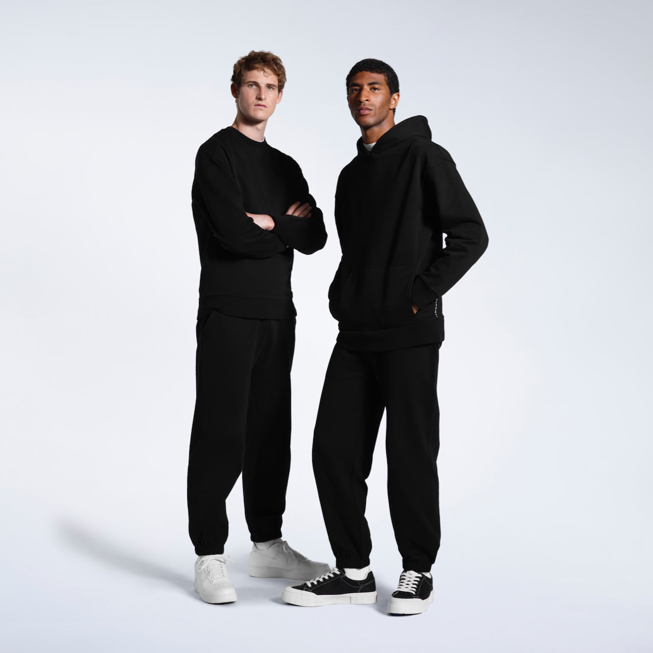 Two men modelling plain black heavyweight, organic cotton tracksuit sets.