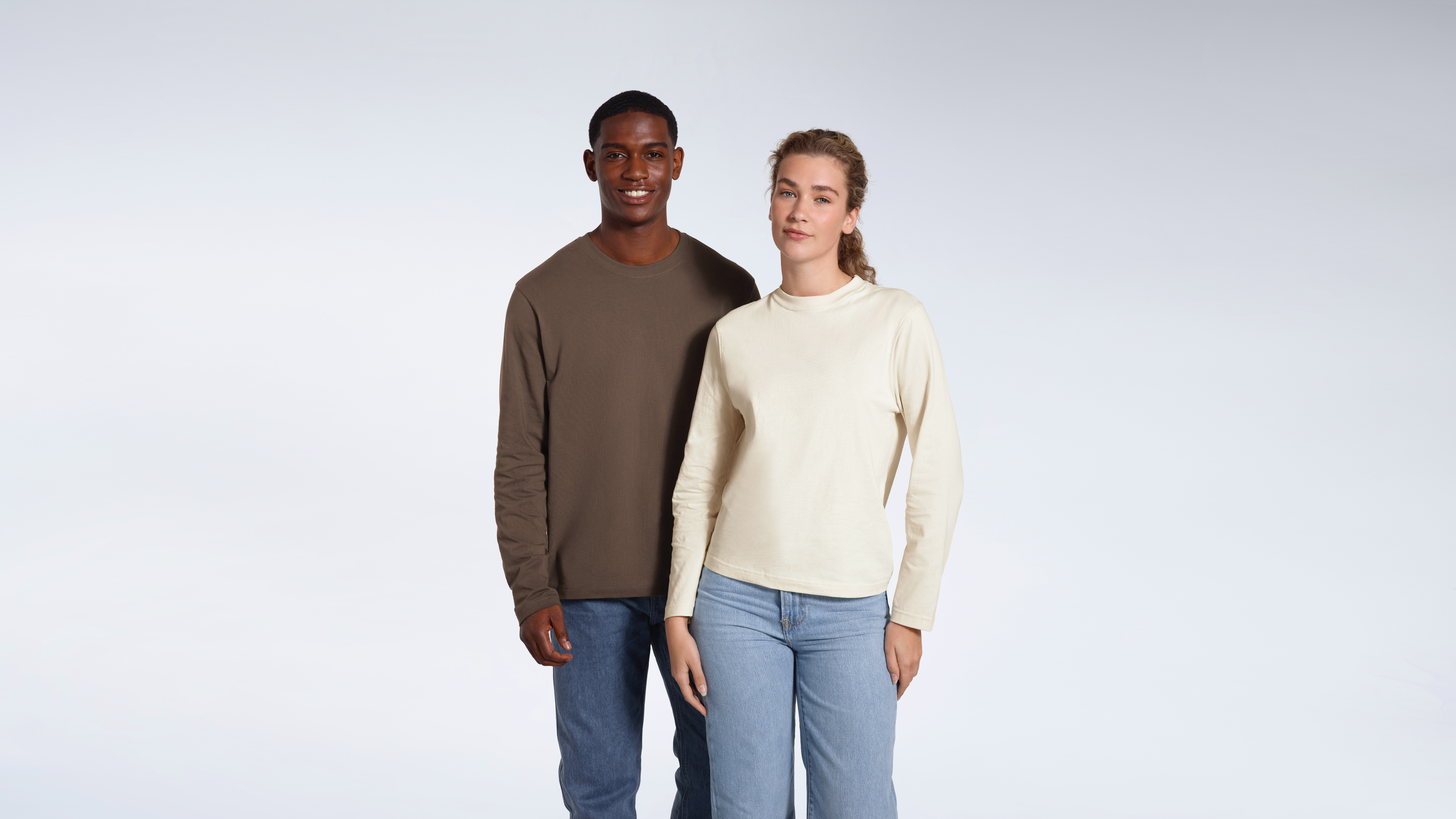 A man and woman smiling into the camera, modelling plain dark khaki and ecru coloured long sleeve t shirts.