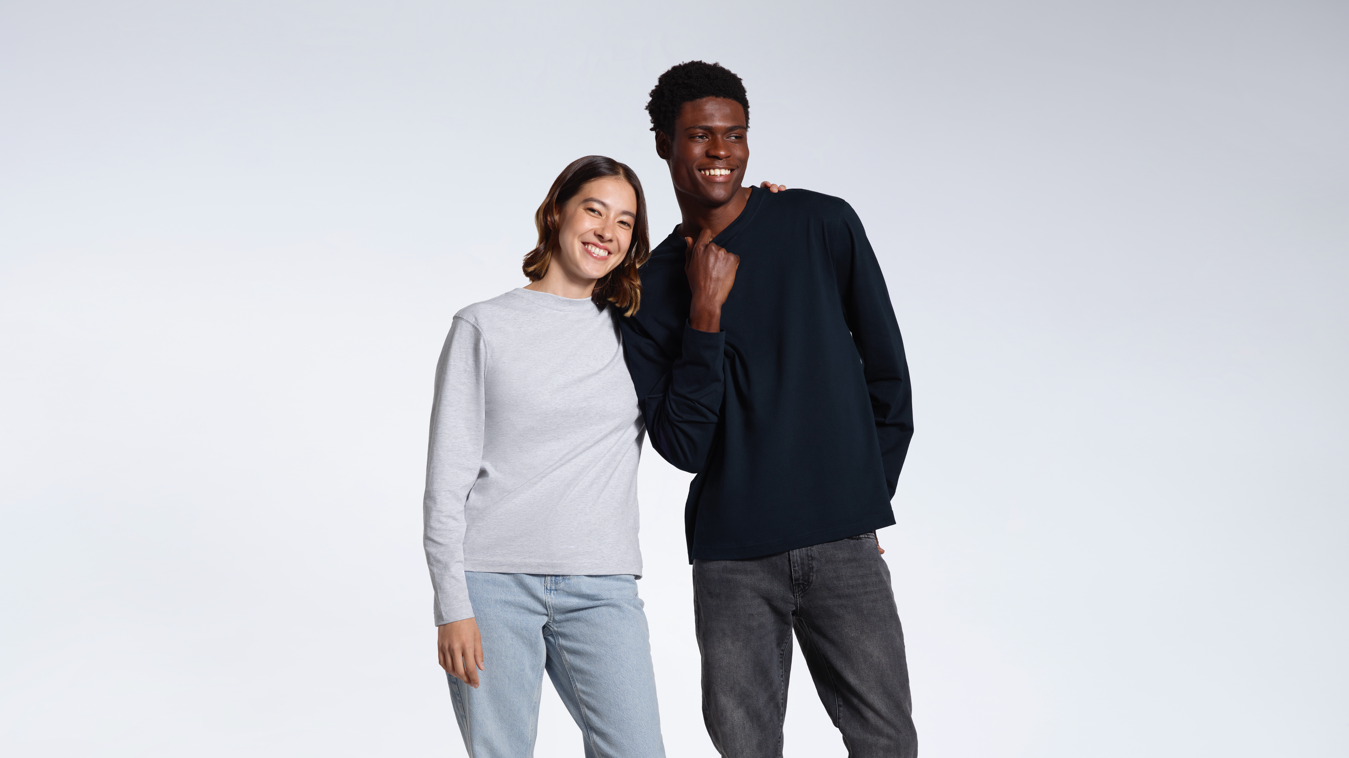 A man and woman smiling into the camera with their arms round eachother, modelling plain grey and navy organic cotton long sleeve t shirts.