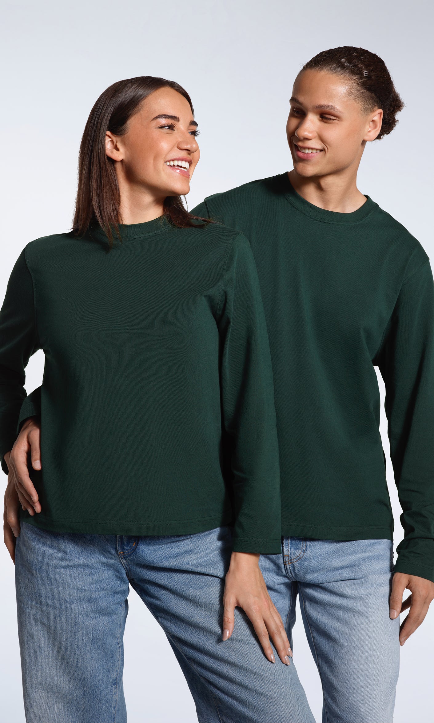 A man with his arm round a woman, both smiling, modelling matching plain green, organic cotton, long sleeve t shirts.
