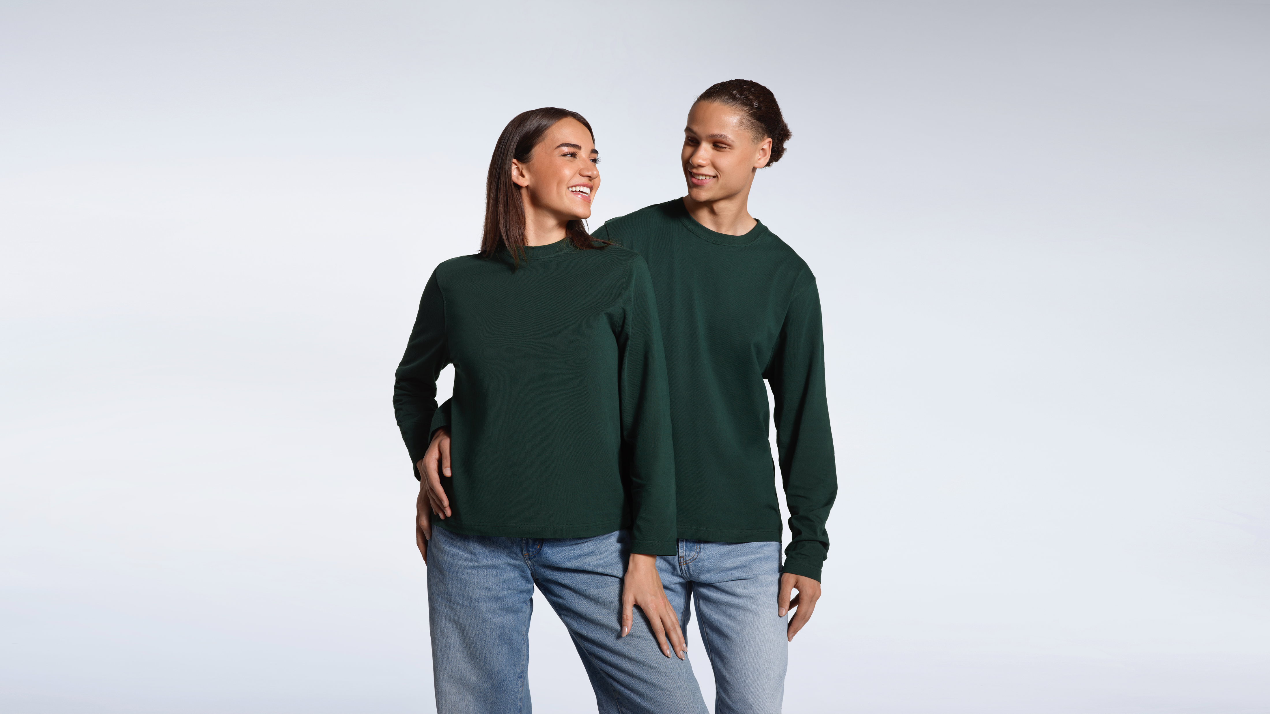 A man with his arm round a woman, both smiling, modelling matching plain green, organic cotton, long sleeve t shirts.