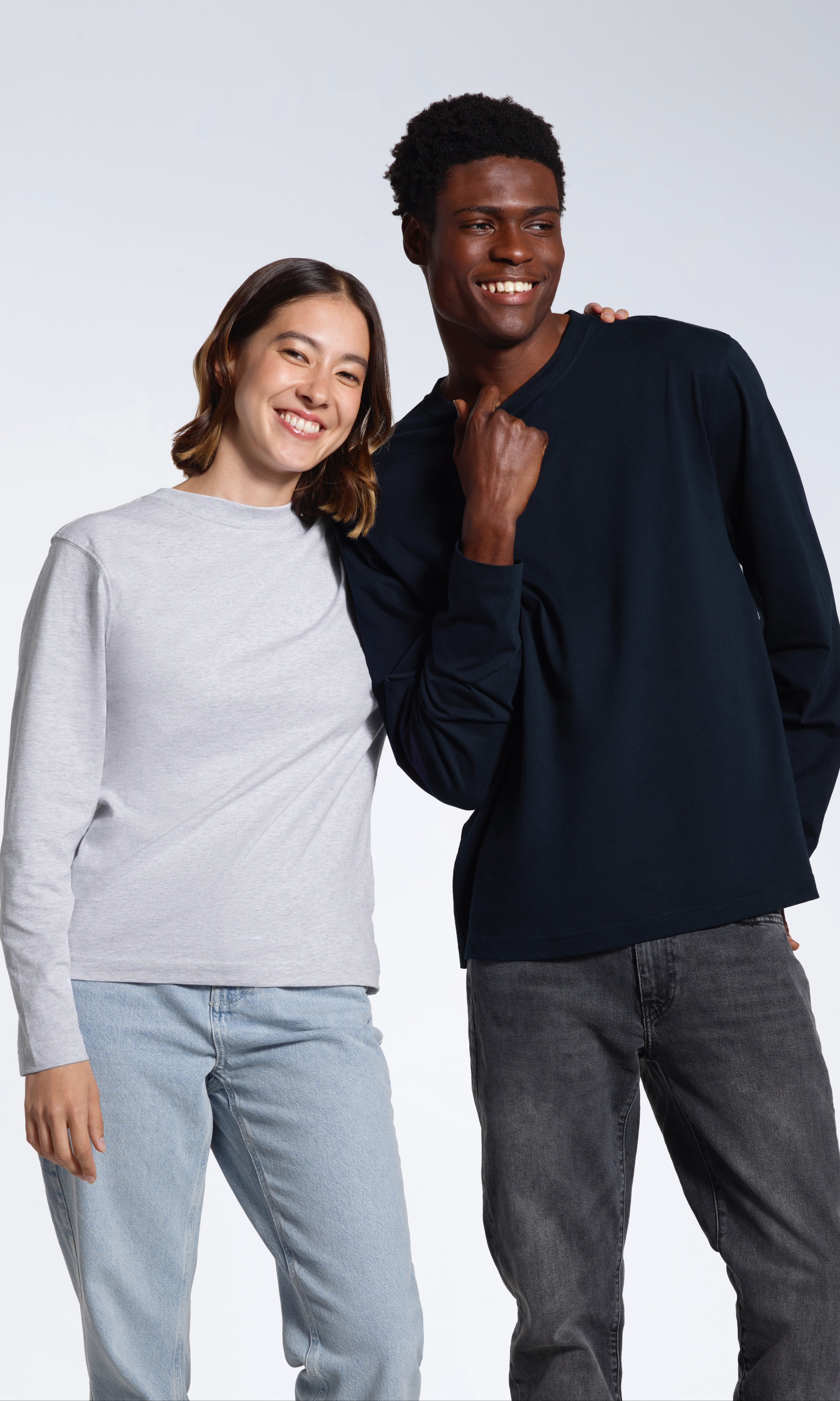 A man and woman smiling into the camera with their arms round eachother, modelling plain grey and navy organic cotton long sleeve t shirts.