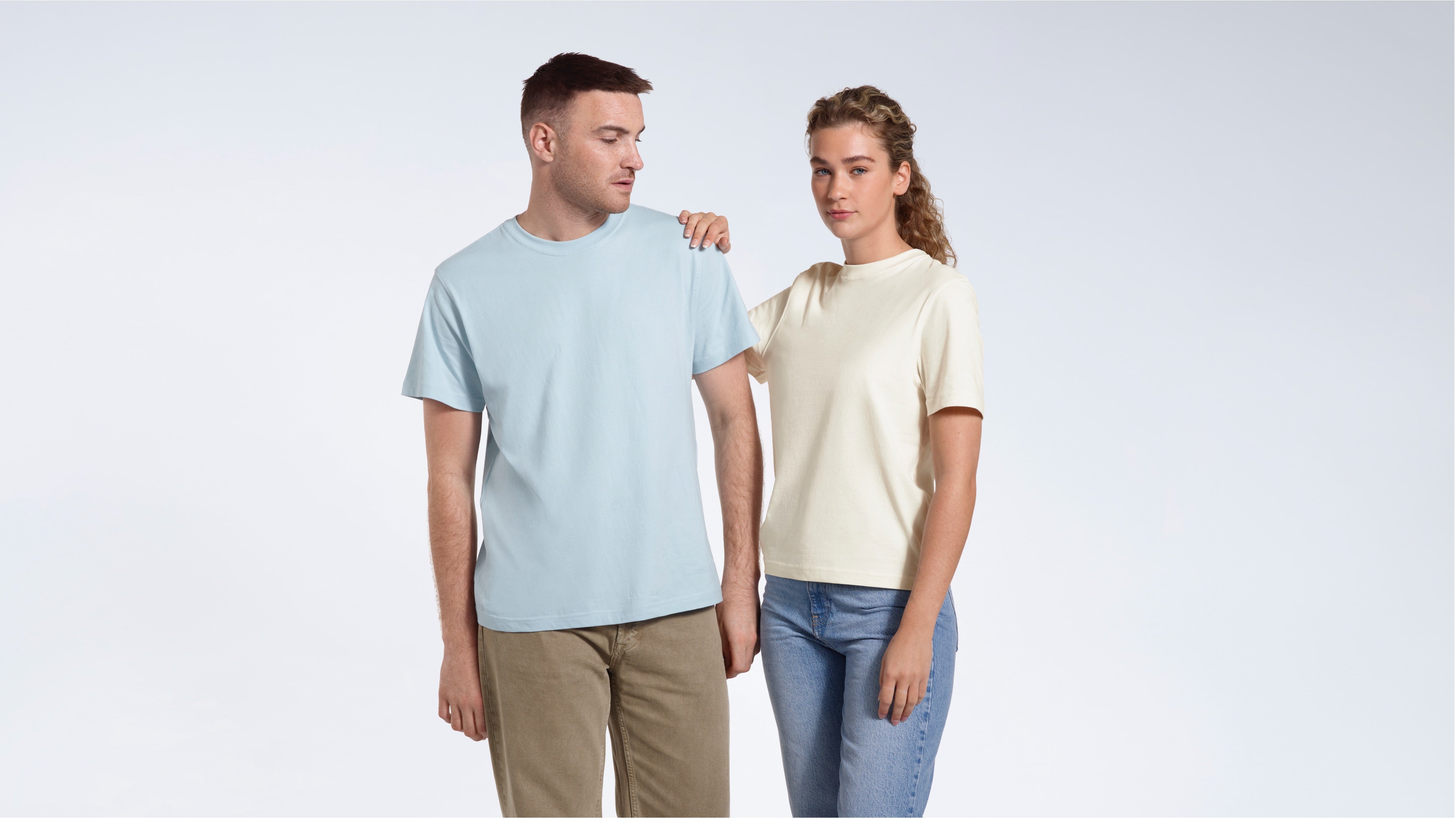 A woman looking into the camera with her hand on the man's shoulder, modelling plain light blue and ecru organic cotton t shirts.