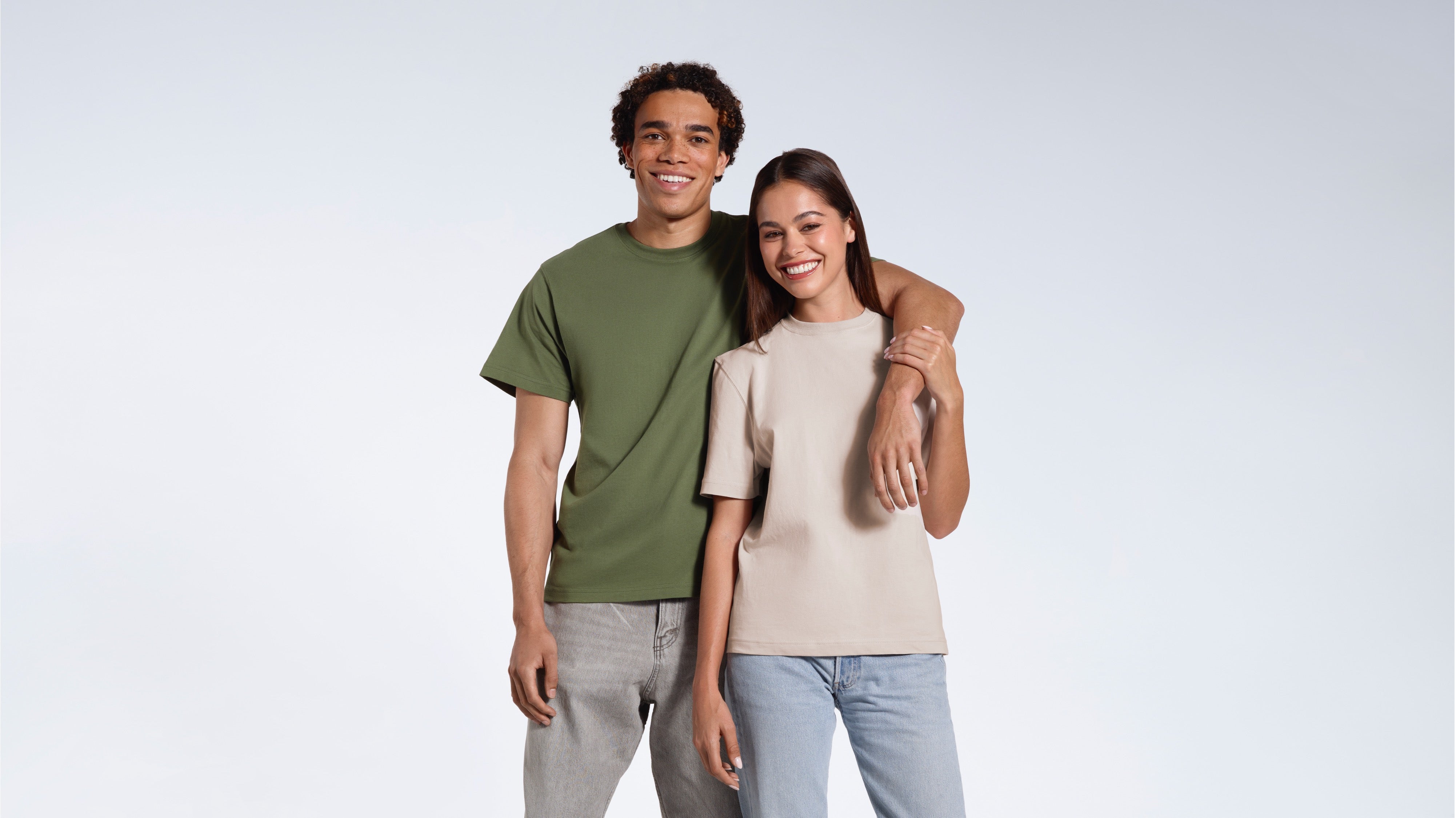 A man with his arm round the woman's shoulder, both smiling into the camera, modelling plain khaki green and stone coloured organic cotton t shirts.