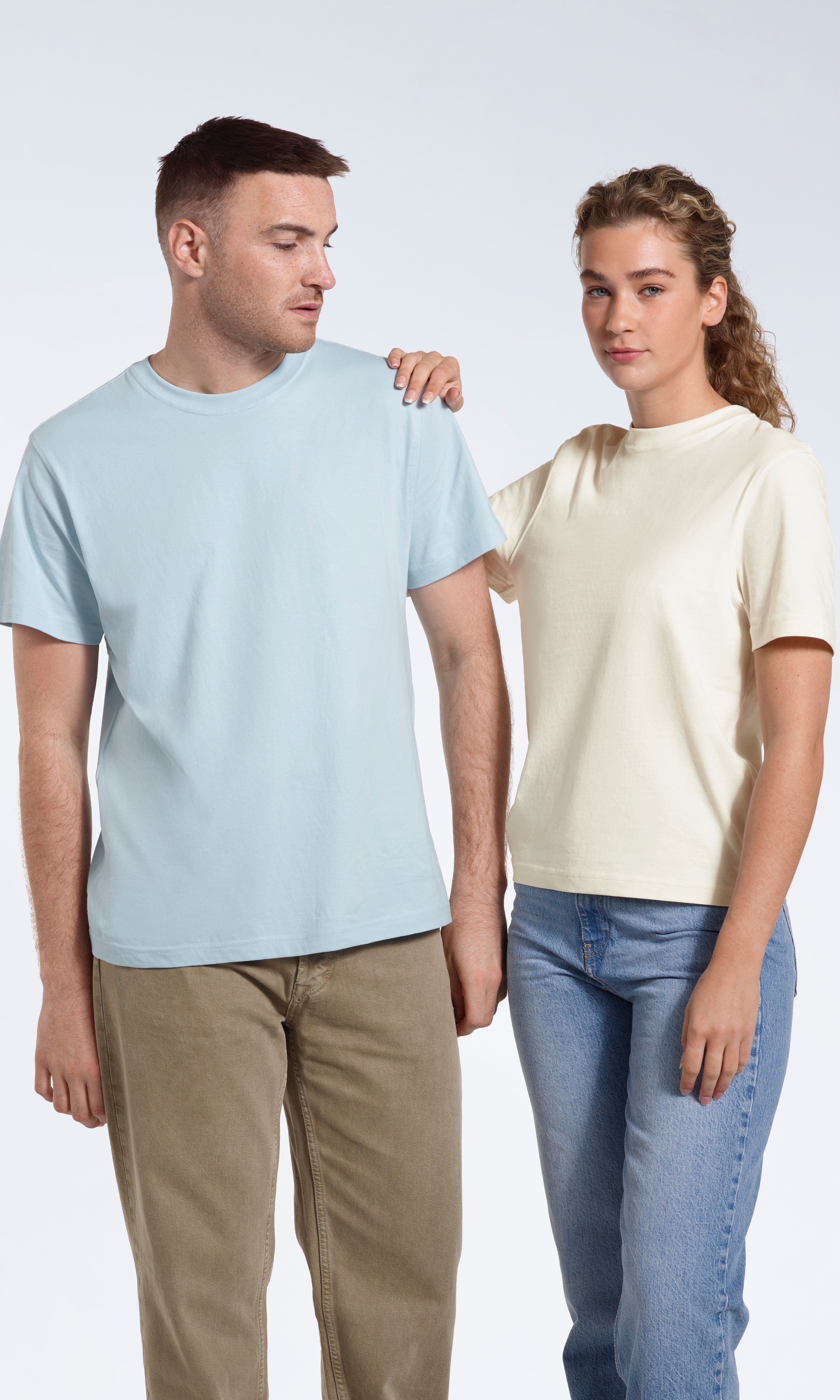A woman looking into the camera with her hand on the man's shoulder, modelling plain light blue and ecru organic cotton t shirts.