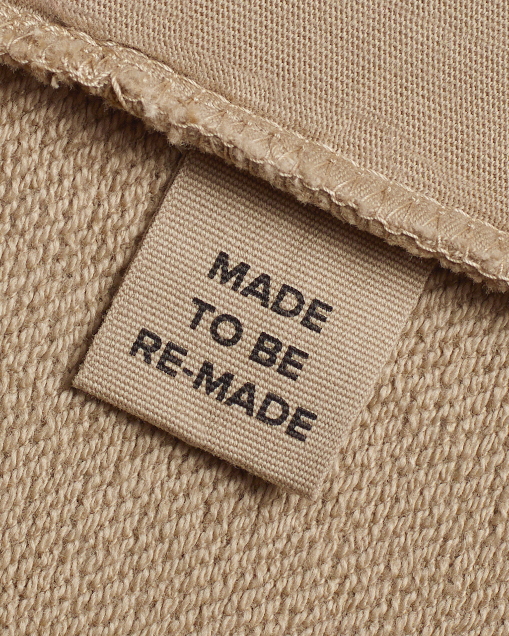 A close up of a label tag in a beige, organic cotton overshirt that says 'made to be re made'.