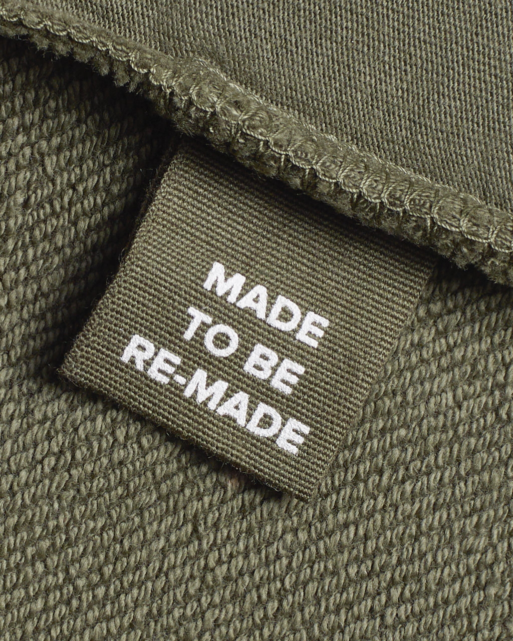 A close up of a label tag in a olive, organic cotton overshirt that says 'made to be re made'.