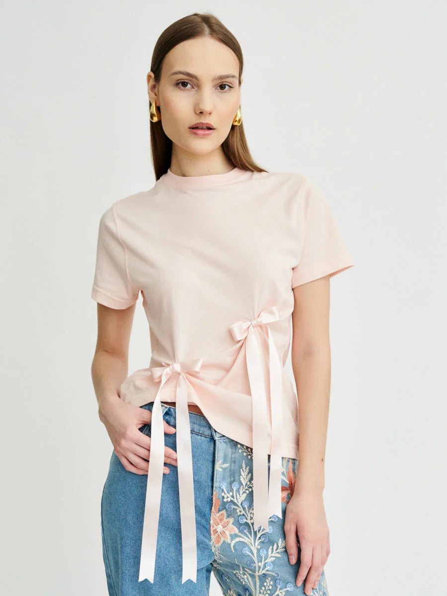 A woman looking into the camera, modelling a pink organic cotton t-shirt with bows attached to it.
