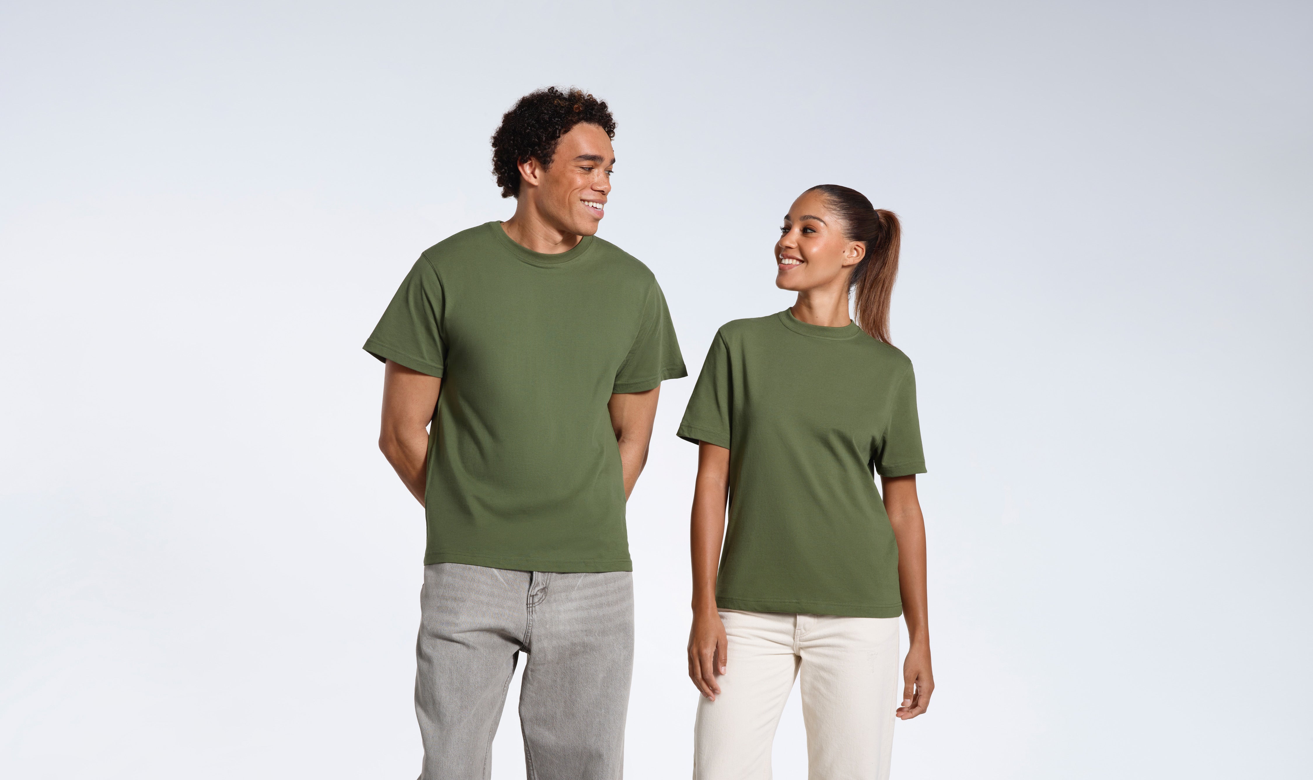 A man and woman looking at eachother smiling, wearing matching khaki green short sleeve organic cotton t shirts.