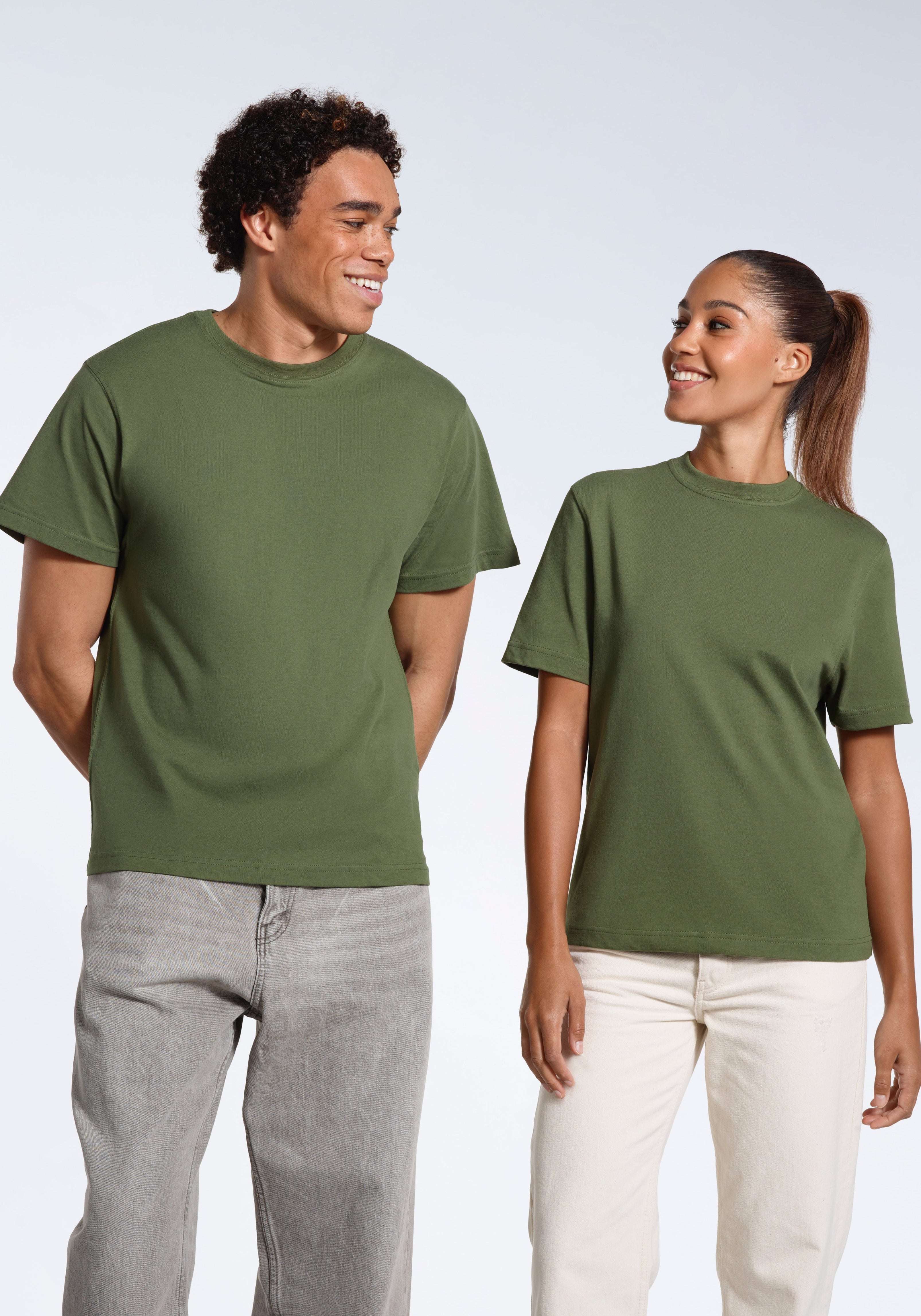 A man and woman looking at eachother smiling, wearing matching khaki green short sleeve organic cotton t shirts.