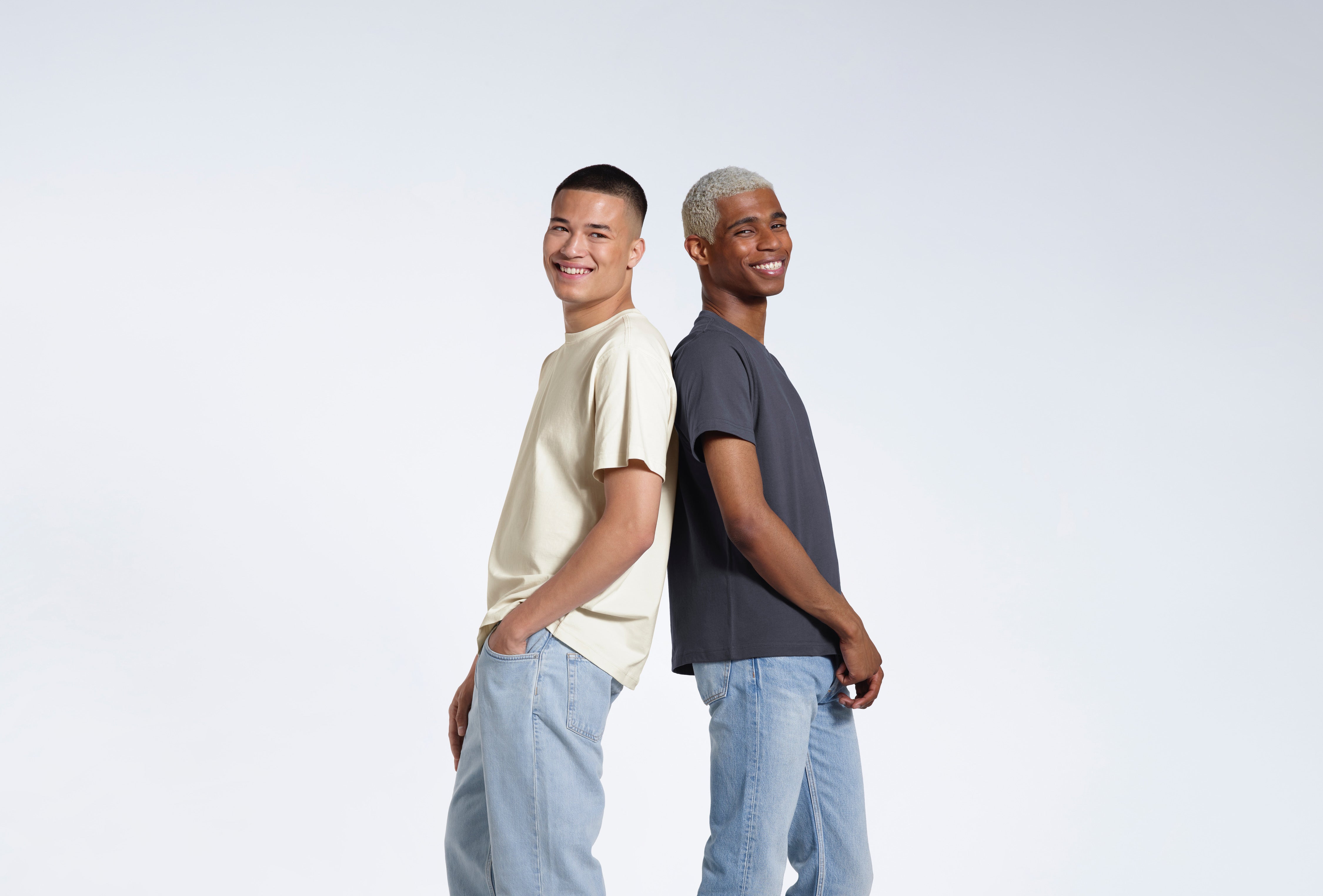 Two men smiling back to back, modelling ecru and dark grey coloured short sleeve organic cotton t shirts.
