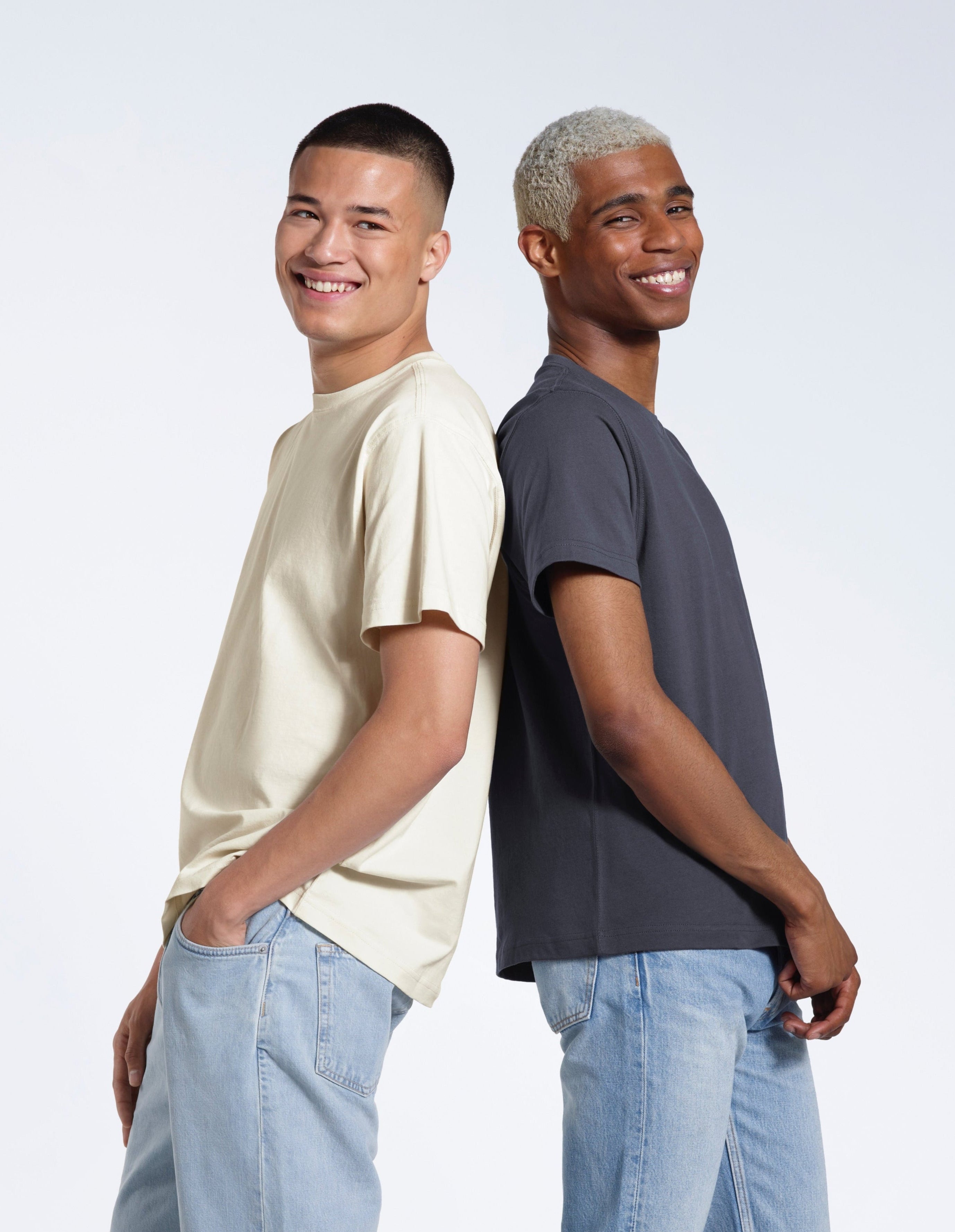 Two men smiling back to back, modelling ecru and dark grey coloured short sleeve organic cotton t shirts.