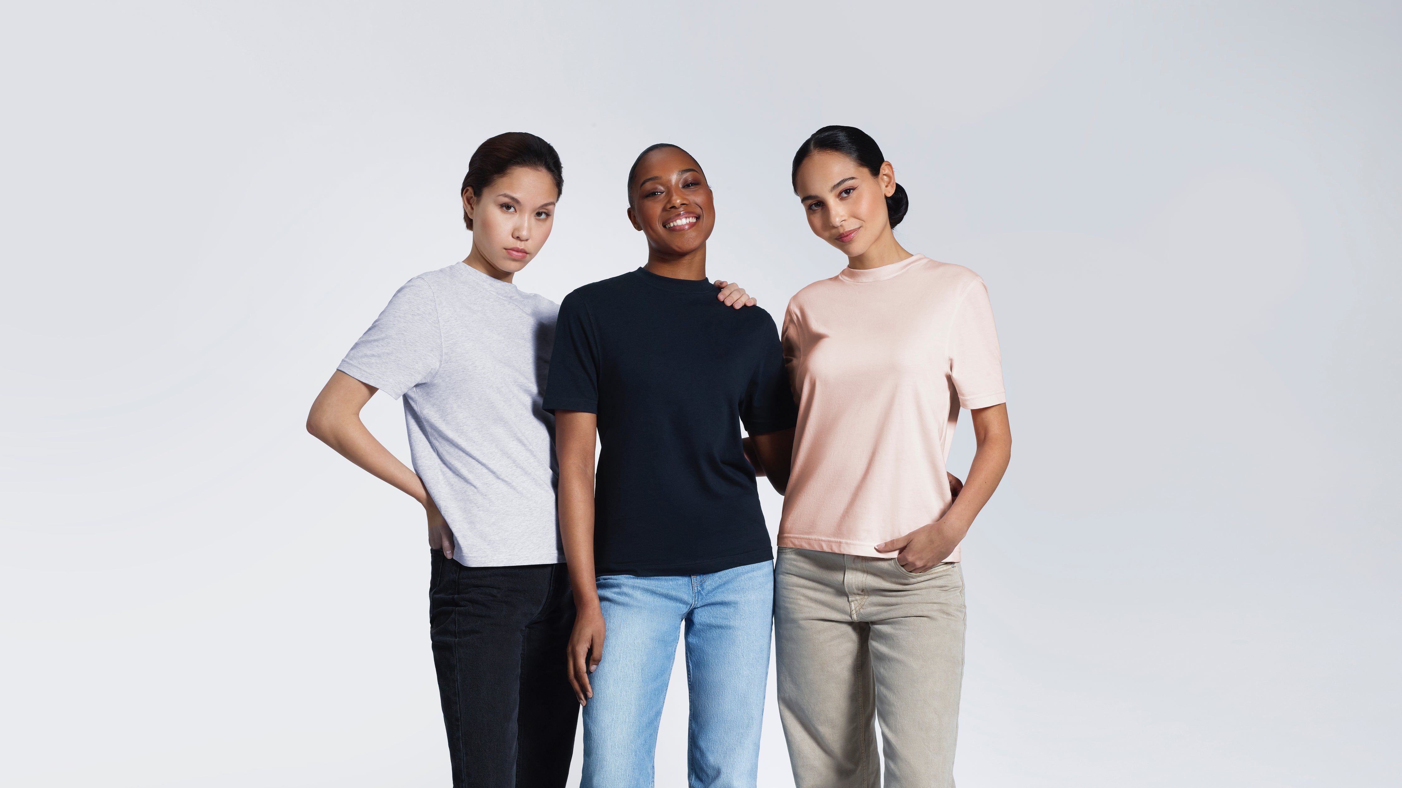 Three women with their arms round each other looking into the camera, modelling plain grey, navy and pink organic cotton t shirts.
