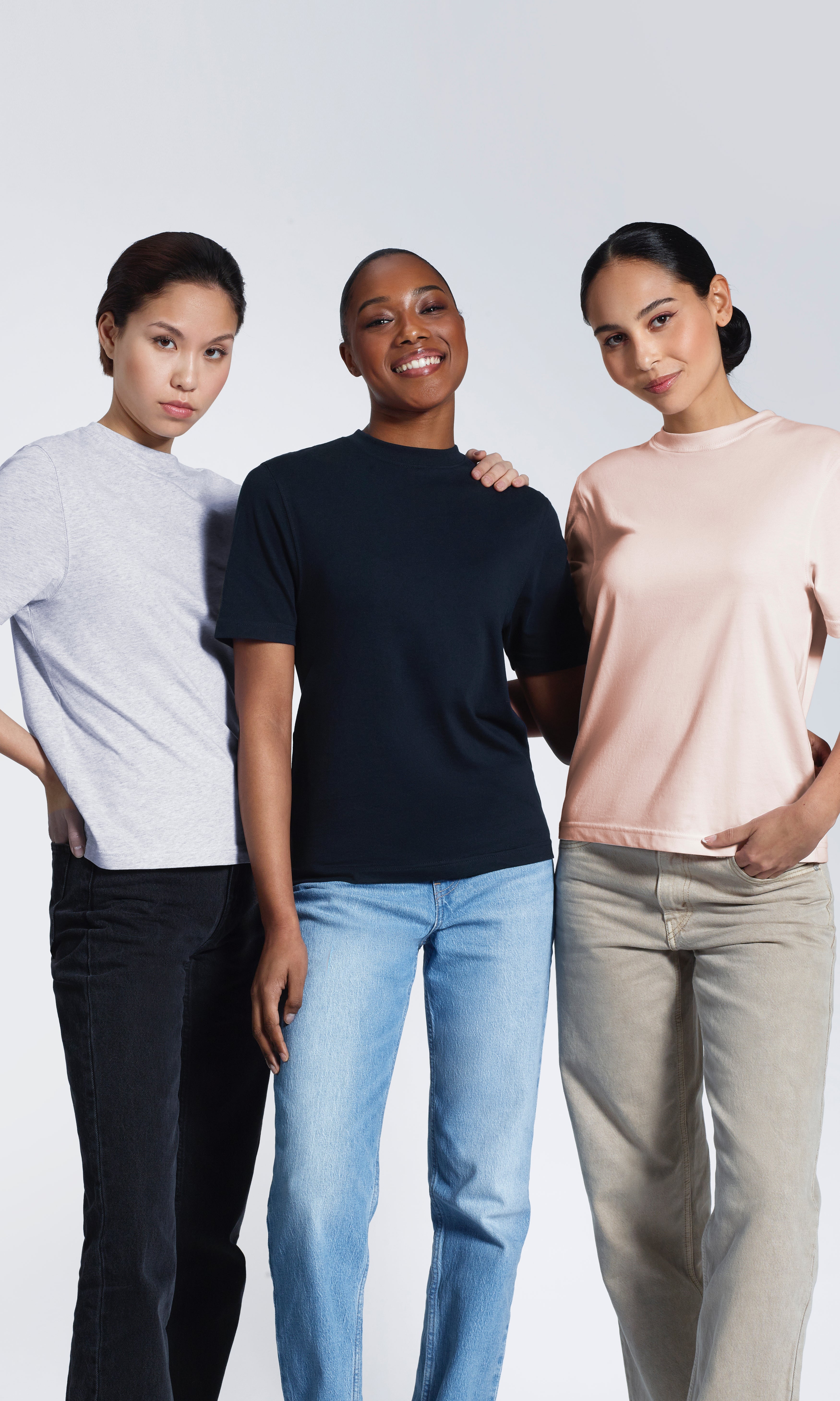 Three women with their arms round each other looking into the camera, modelling plain grey, navy and pink organic cotton t shirts.