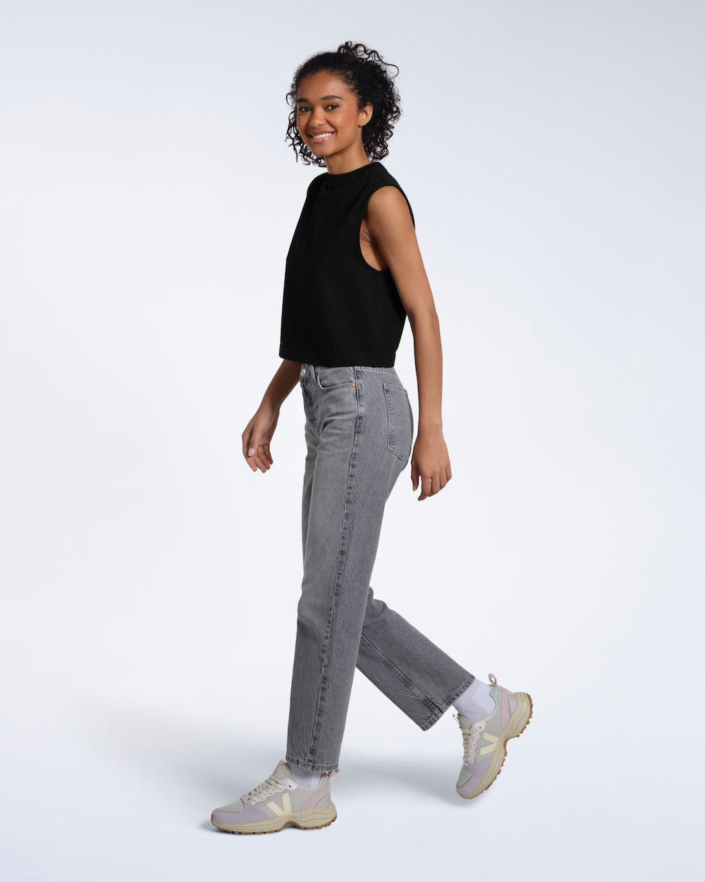 A full body photo of a woman walking and smiling, modelling a plain black sleeveless organic cotton tank top.