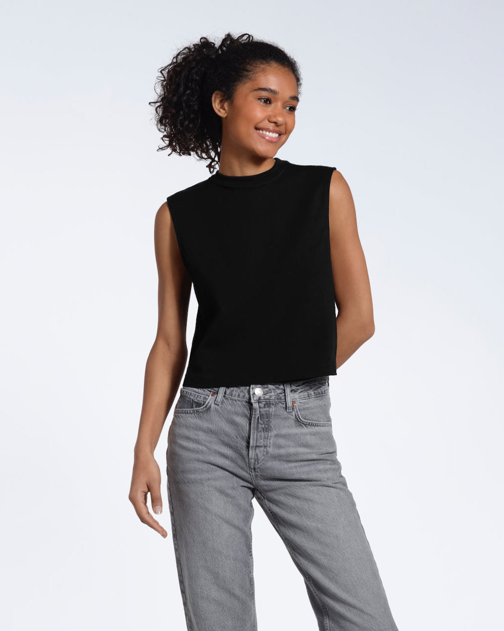 A woman smiling with one arm behind her, modelling a plain black sleeveless organic cotton tank top.