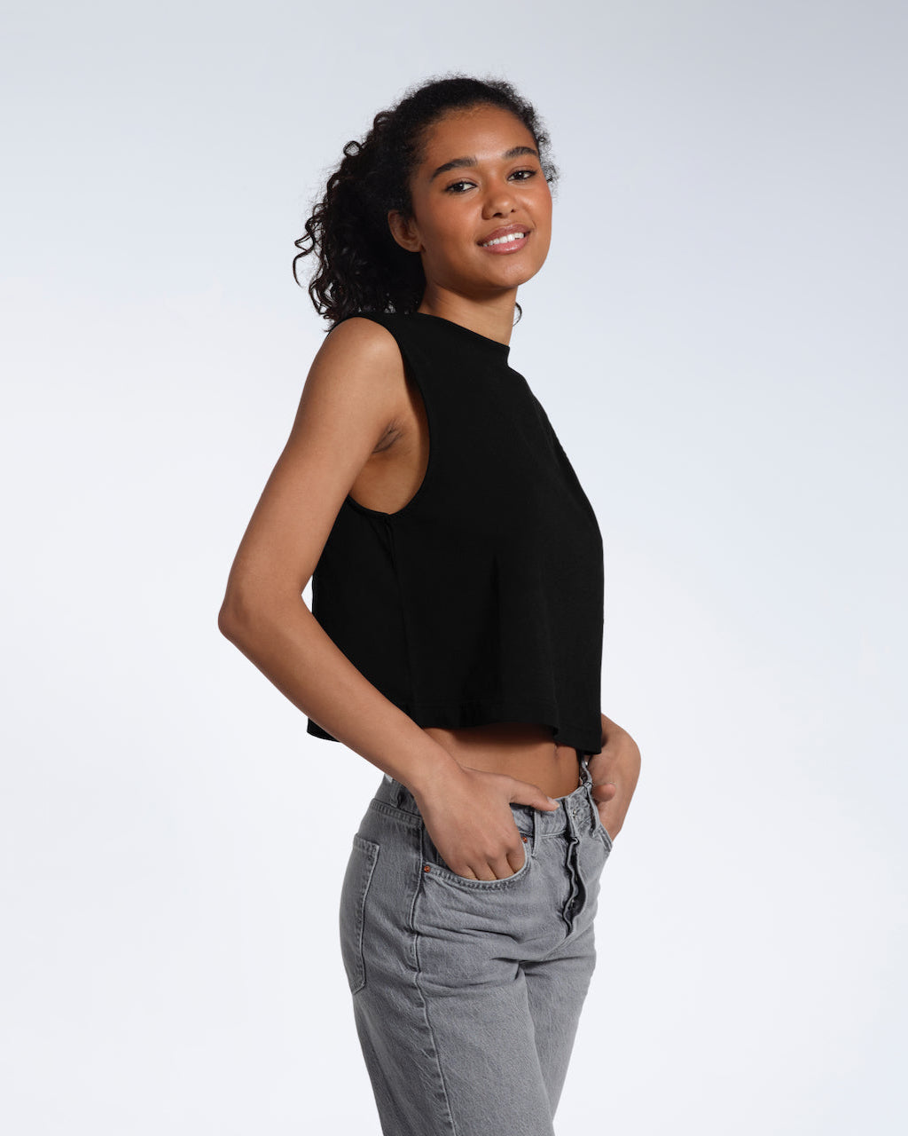 A woman smiling at the camera with her hands in her jeans pockets, modelling a plain black sleeveless organic cotton tank top.