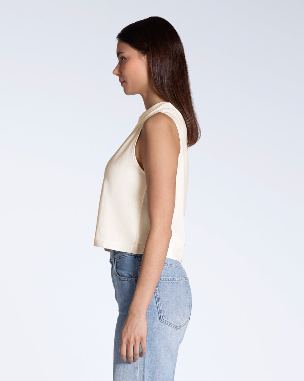 A woman facing side on, modelling a plain natural coloured, organic cotton, crew neck tank top.