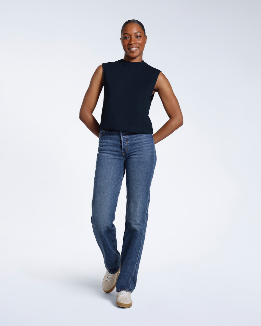 A full body photo of a woman smiling with her arms behind her back, modelling a plain navy sleeveless organic cotton tank top.