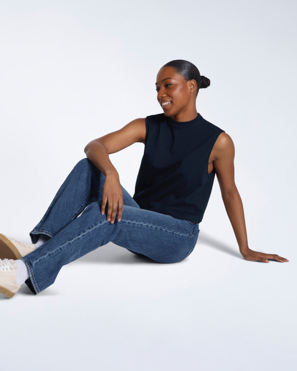 A woman sitting on the floor smiling, modelling a plain navy sleeveless organic cotton tank top.