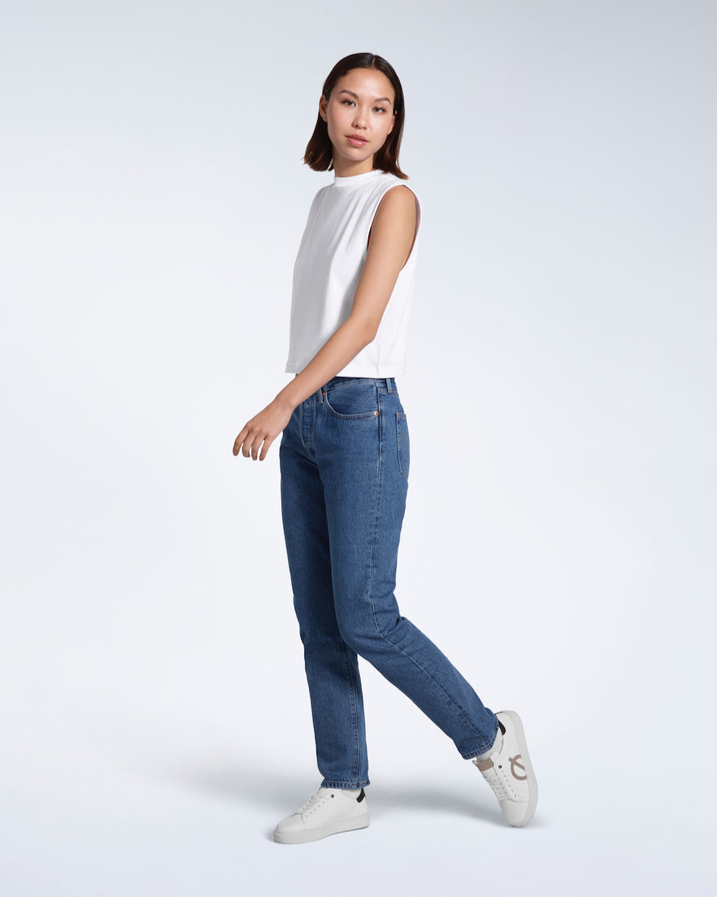 A full body photo of a woman walking, modelling a plain white sleeveless organic cotton tank top.