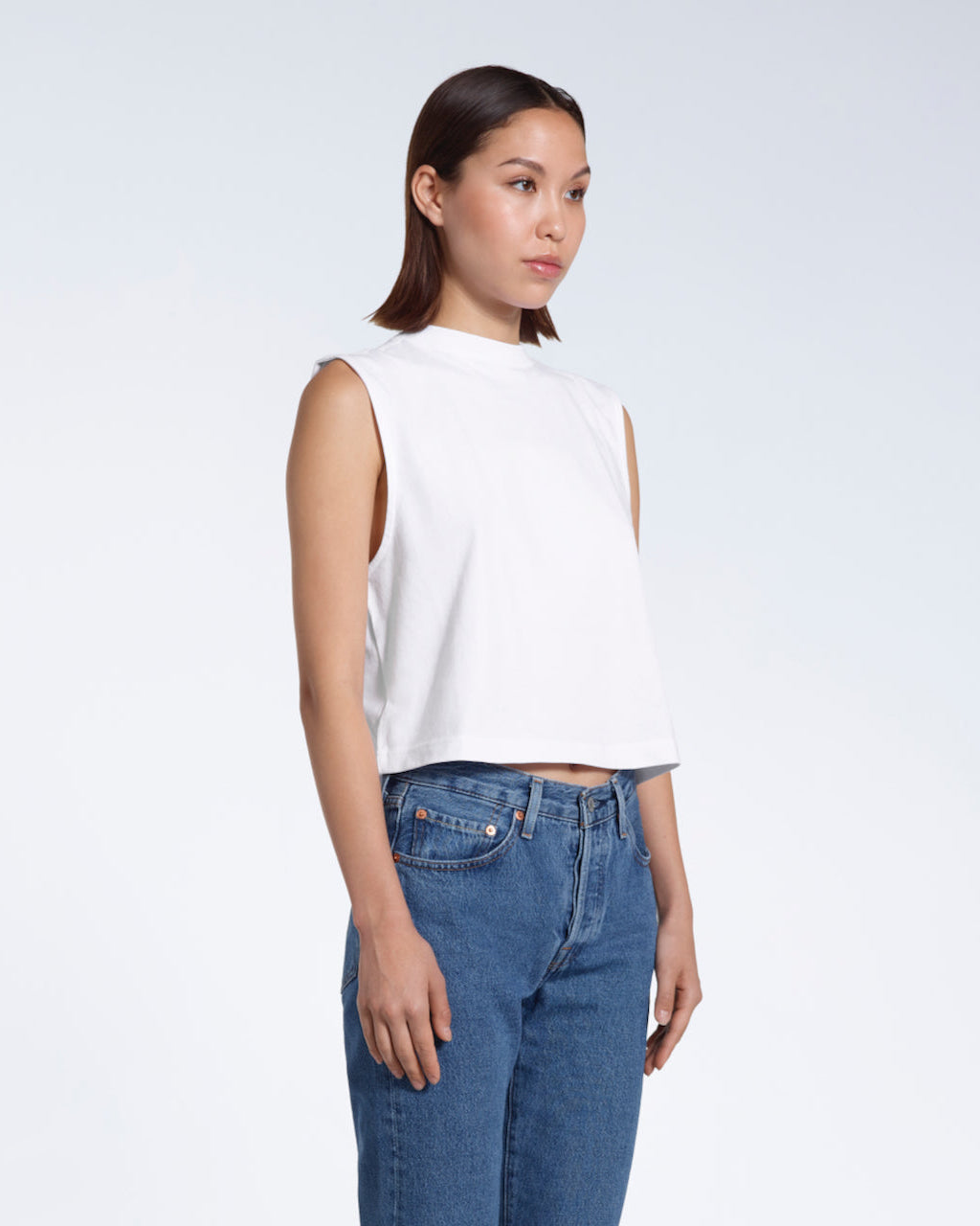 A woman at an angle with her hands by her sides, modelling a plain white sleeveless organic cotton tank top.