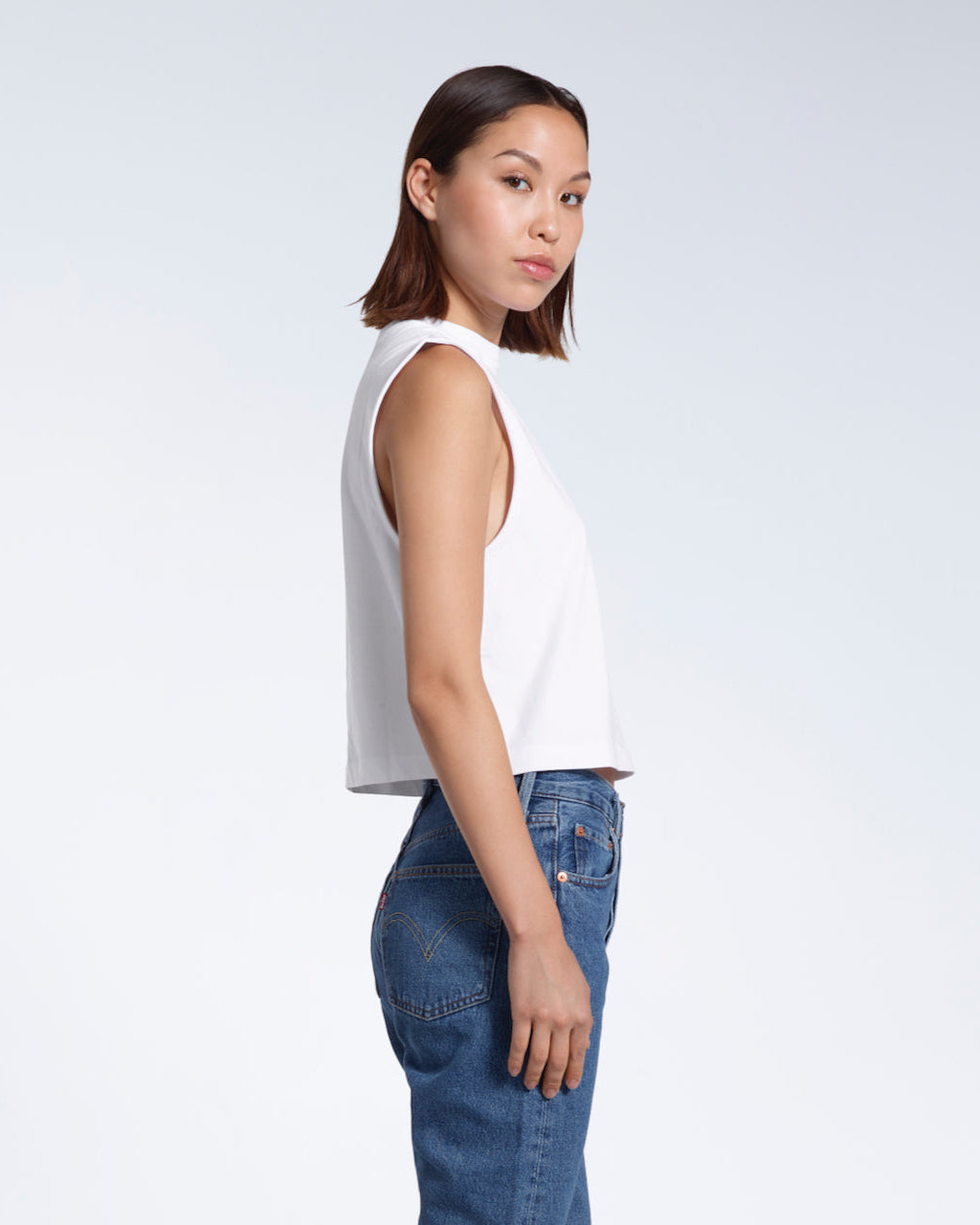 A woman facing side on and looking at the camera, modelling a plain white sleeveless organic cotton tank top.