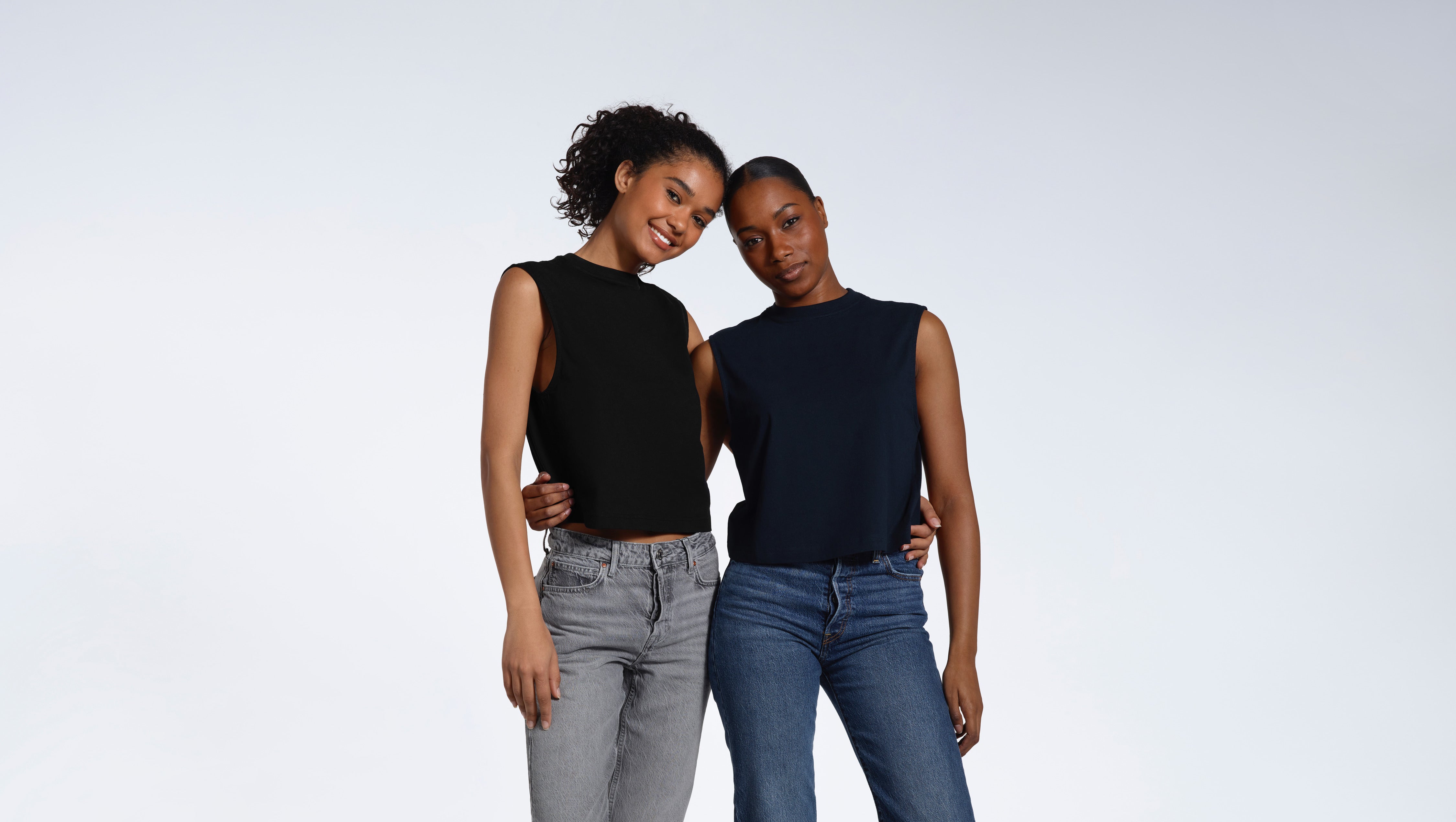 Two women with their arms around eachother smiling into the camera, modelling plain black and navy organic cotton tank tops.