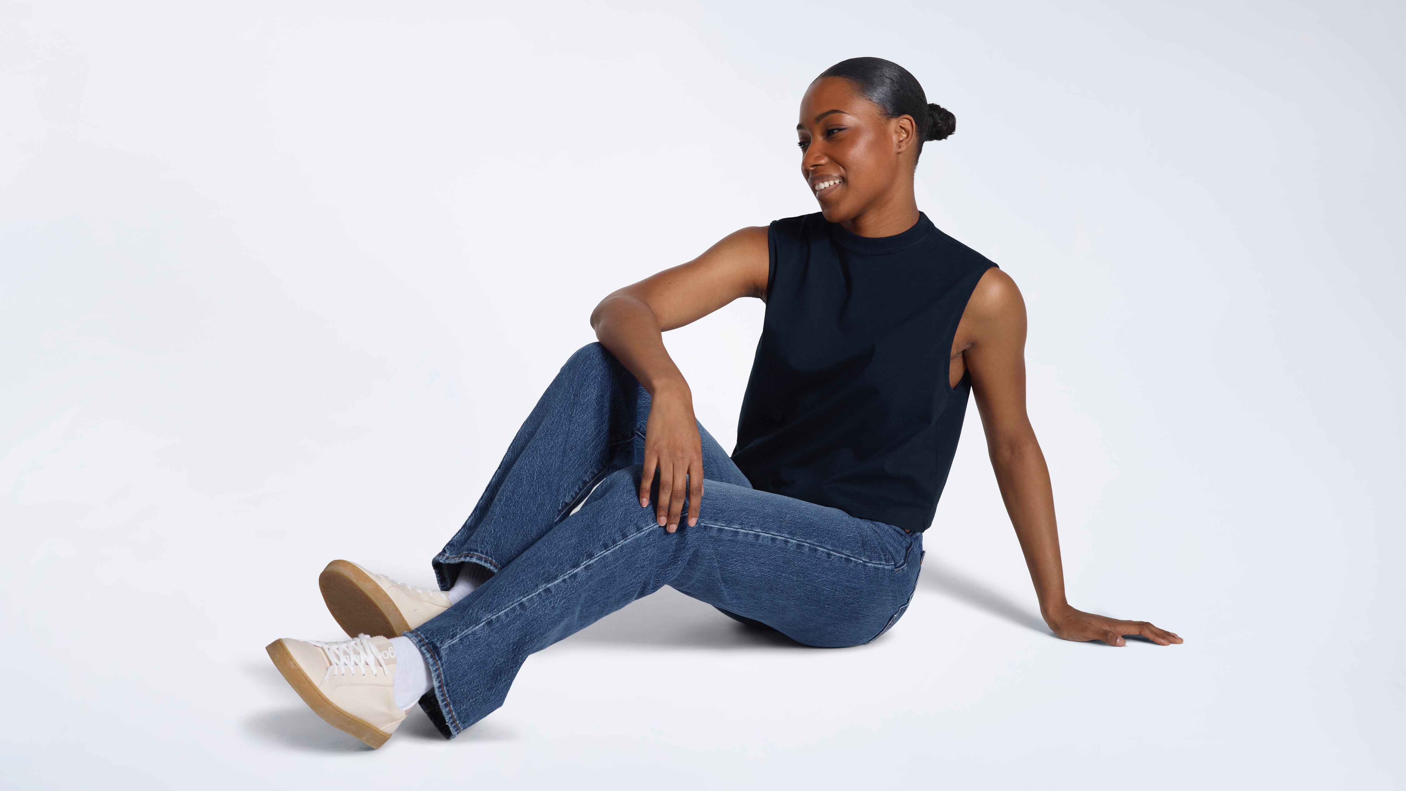 A woman sitting on the floor smiling, modelling a plain navy sleeveless organic cotton tank top.