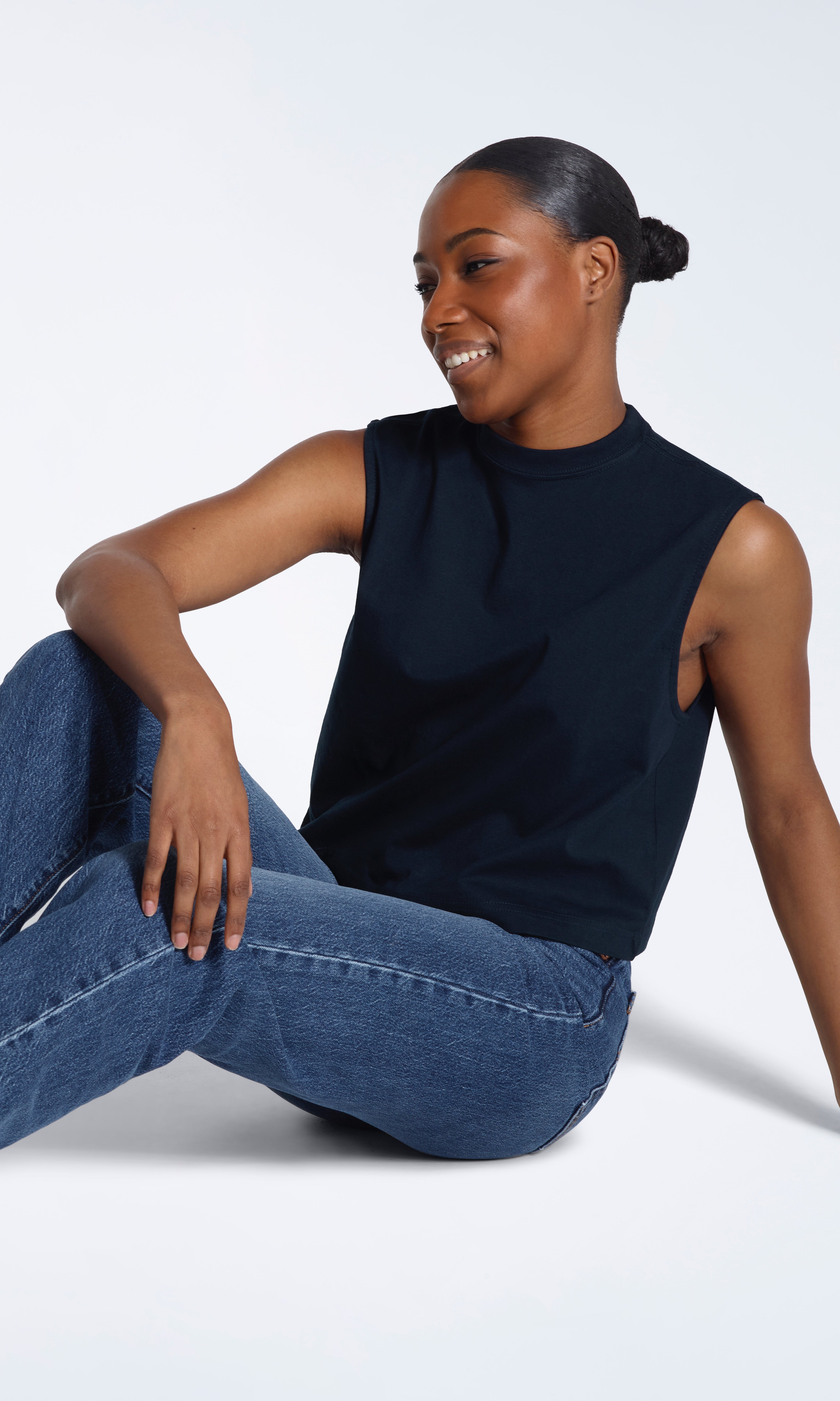 A woman sitting on the floor smiling, modelling a plain navy sleeveless organic cotton tank top.