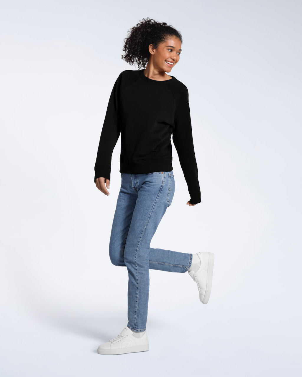 A full body photo of a woman smiling with one leg bent, modelling a plain black french terry organic cotton sweatshirt.
