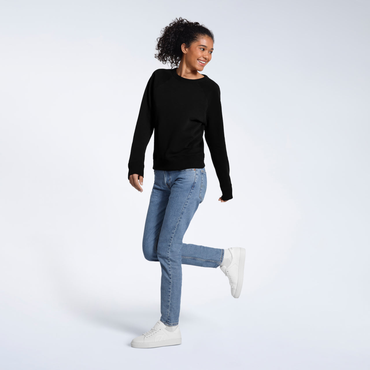A full body photo of a woman smiling with one leg bent, modelling a plain black french terry organic cotton sweatshirt.