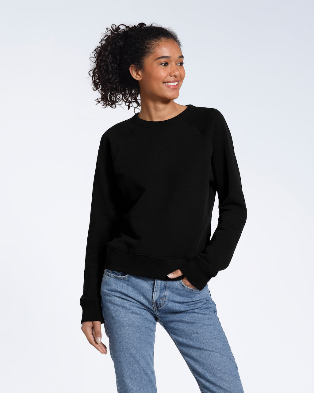 A woman looking away from the camera smiling with a hand in her pocket, modelling a plain black french terry organic cotton sweatshirt.