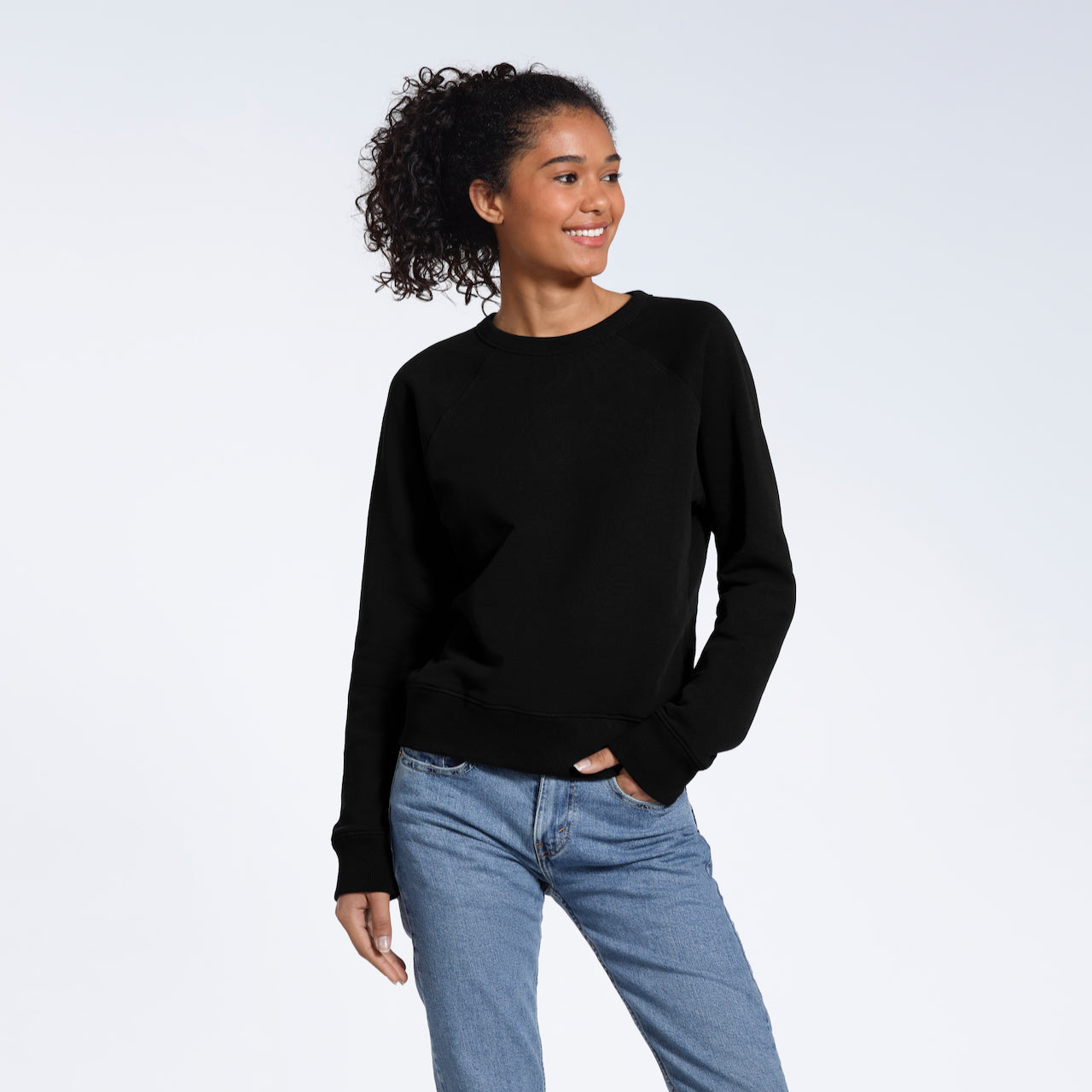 A woman looking away from the camera smiling with a hand in her pocket, modelling a plain black french terry organic cotton sweatshirt.