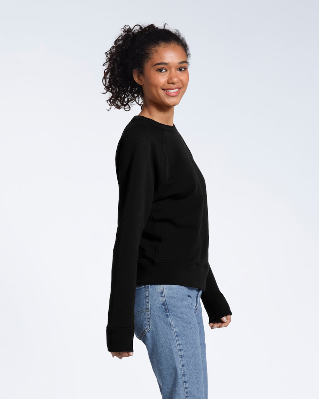 A woman facing side on looking at the camera smiling, modelling a plain black french terry organic cotton sweatshirt.